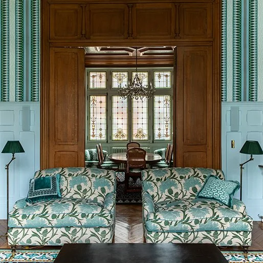 Interieur van een woonkamer met twee gestreepte banken, groene kussens, groene wandlampen, grote houten deur en een eettafel met stoelen, grote raampartij met glas-in-lood en een kroonluchter.