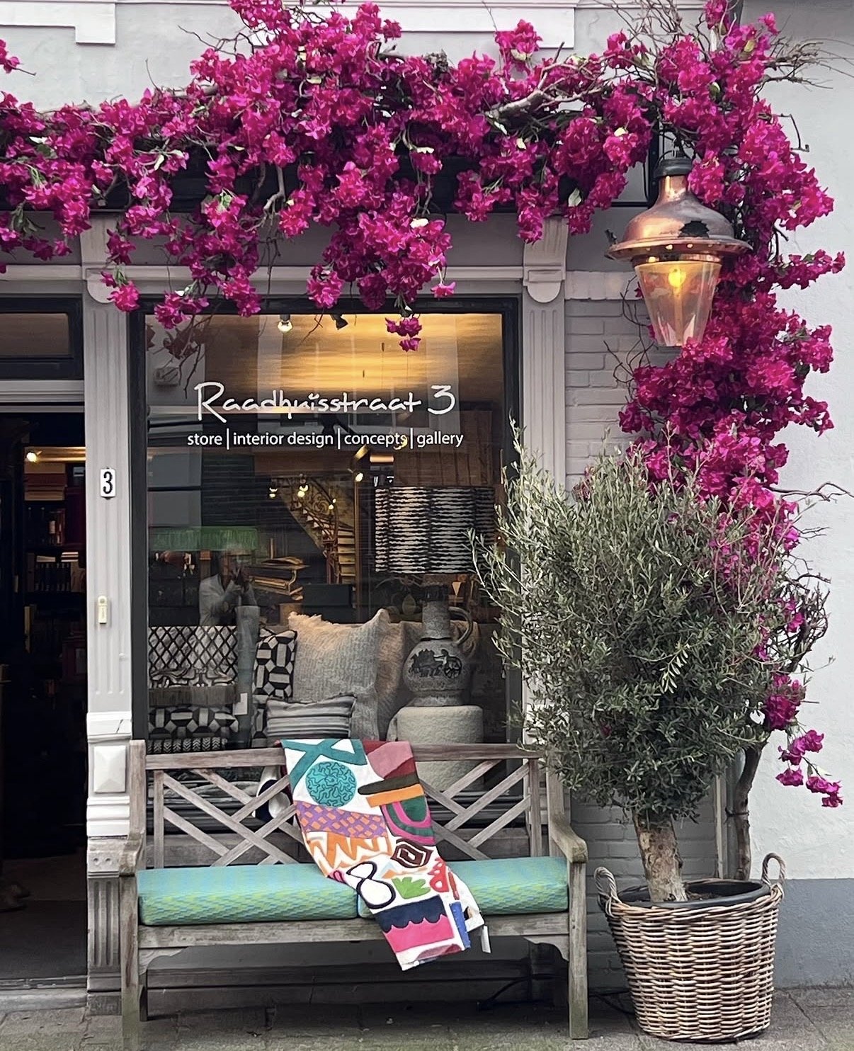 Uitzicht op een winkel met de naam Raadhuisstraat 3, versierd met roze bloemen en een buitenbank met een kleurrijk handdoek of kussen. Binnenshuis zijn meubels en decoraties zichtbaar.