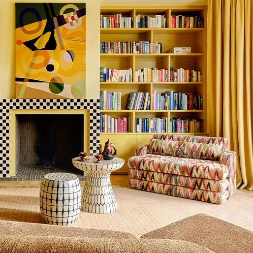 Een woonkamer met een gele boekenplank, een kleurrijke bank, een haard, en twee bijzettafels.