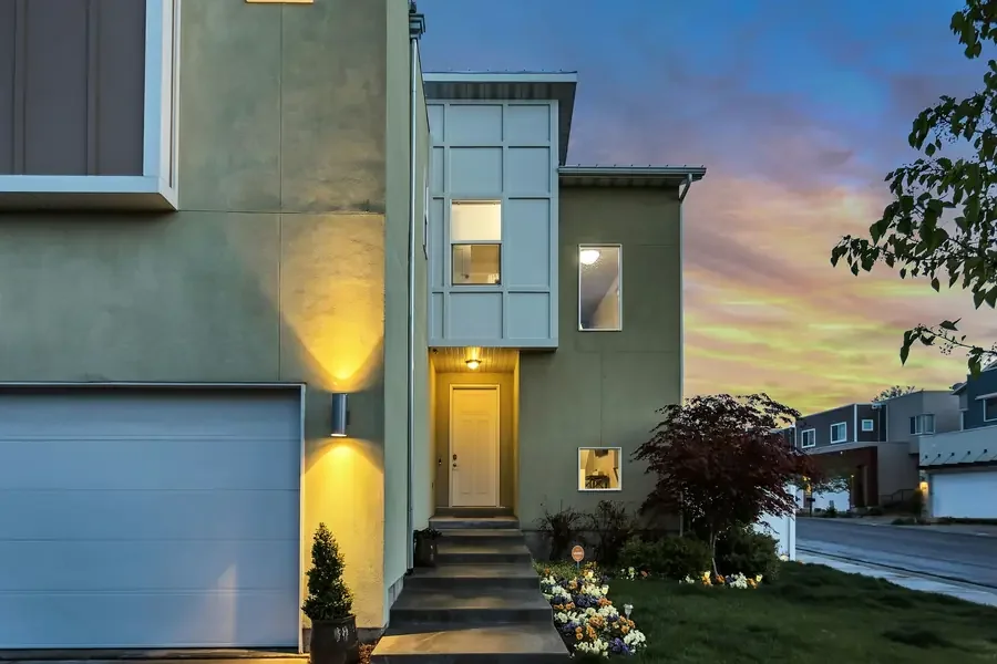 Modern multi-story house with a yellow front door, located on a suburban street during sunset. Exterior features include a beige facade, a garage, illuminated wall-mounted light fixtures, and landscaped front yard with potted plants and flowerbeds.