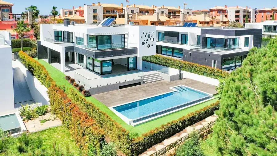 Modern multi-story house with large windows, outdoor pool, wooden deck, and landscaped garden surrounded by colorful hedges and trees in a suburban area.