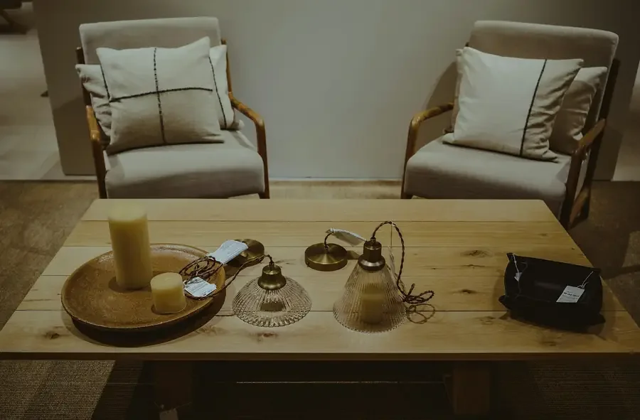 A wooden coffee table with decorative candles and lamp fittings, flanked by two beige armchairs with cushions, in a cozy living room setting.
