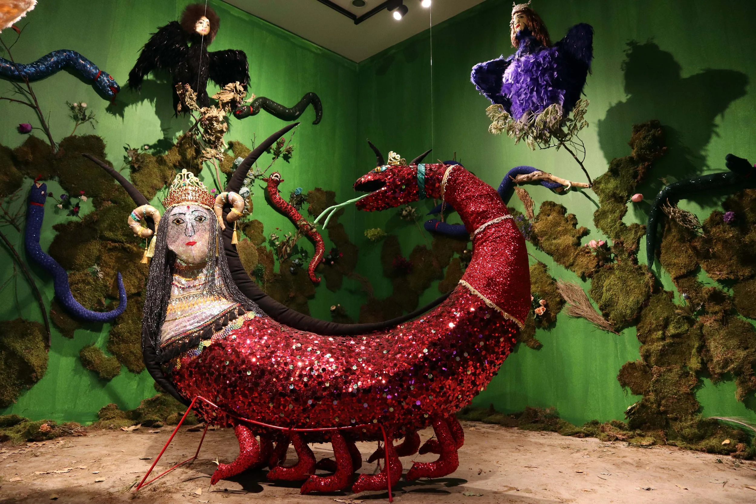 Colorful art installation depicting a mythical creature with a human face and a scorpion body, surrounded by snake-like forms and feathered figures against a green backdrop.