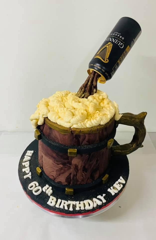 A creative birthday cake designed to look like a beer mug with frothy topping, with an angled Guinness can appearing to pour into it. The cake sits on a base with the text "Happy 60th Birthday Kev."