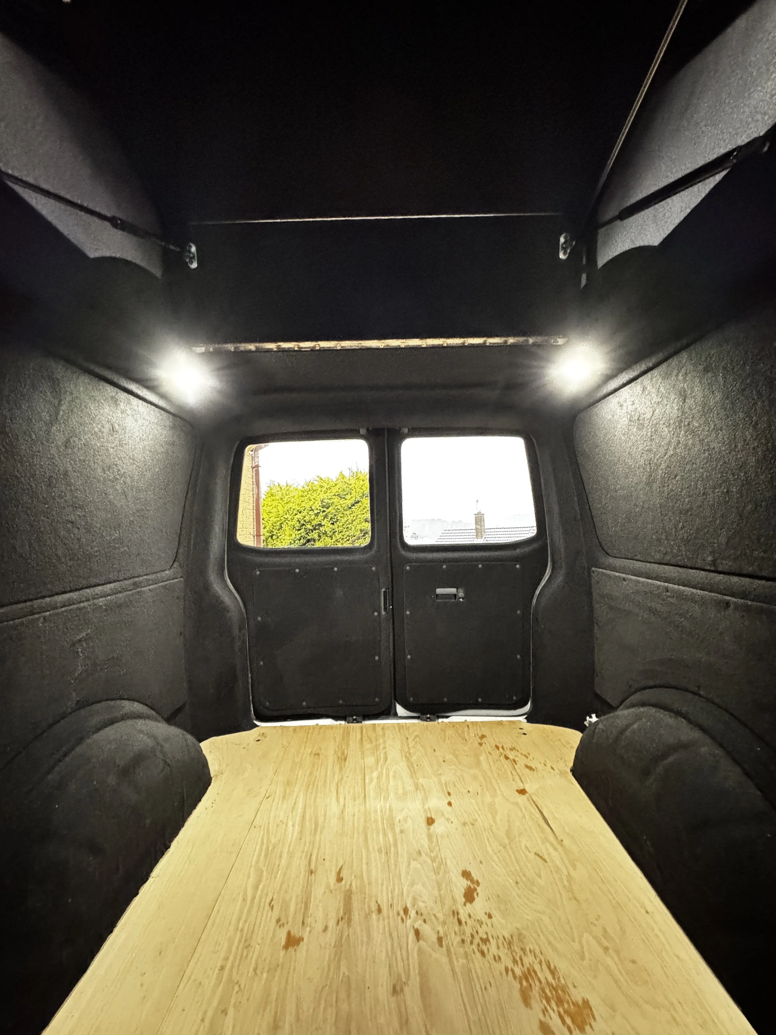 Interior of an empty van with a wooden floor and black walls, featuring two rear doors and overhead lights.