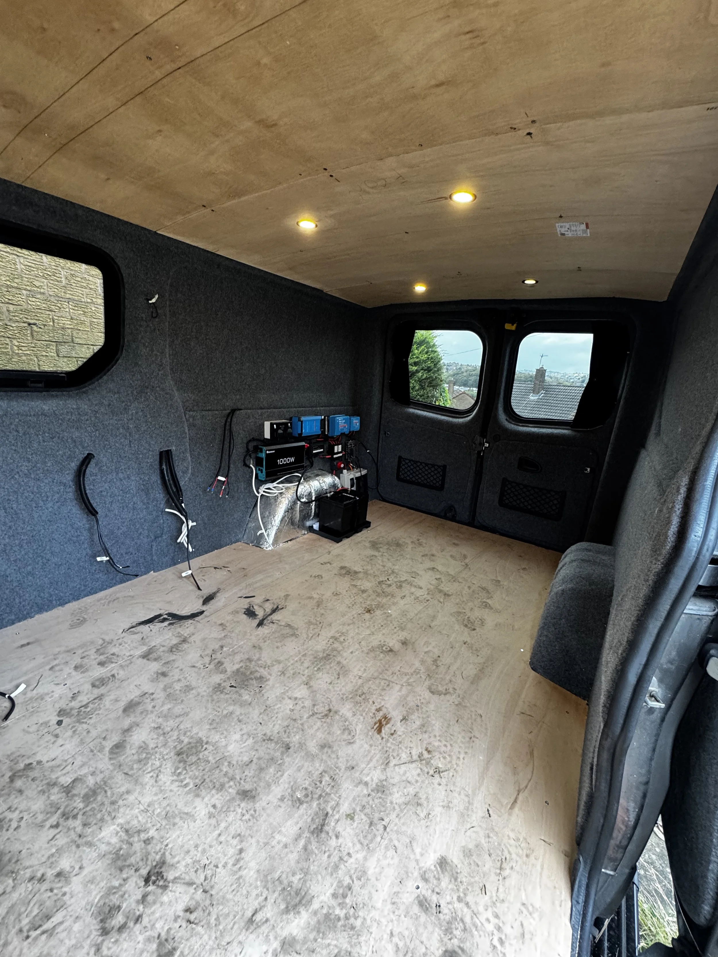 Empty van interior with wooden ceiling, gray carpeted walls, and plywood floor; electrical components and wiring visible on the wall.
