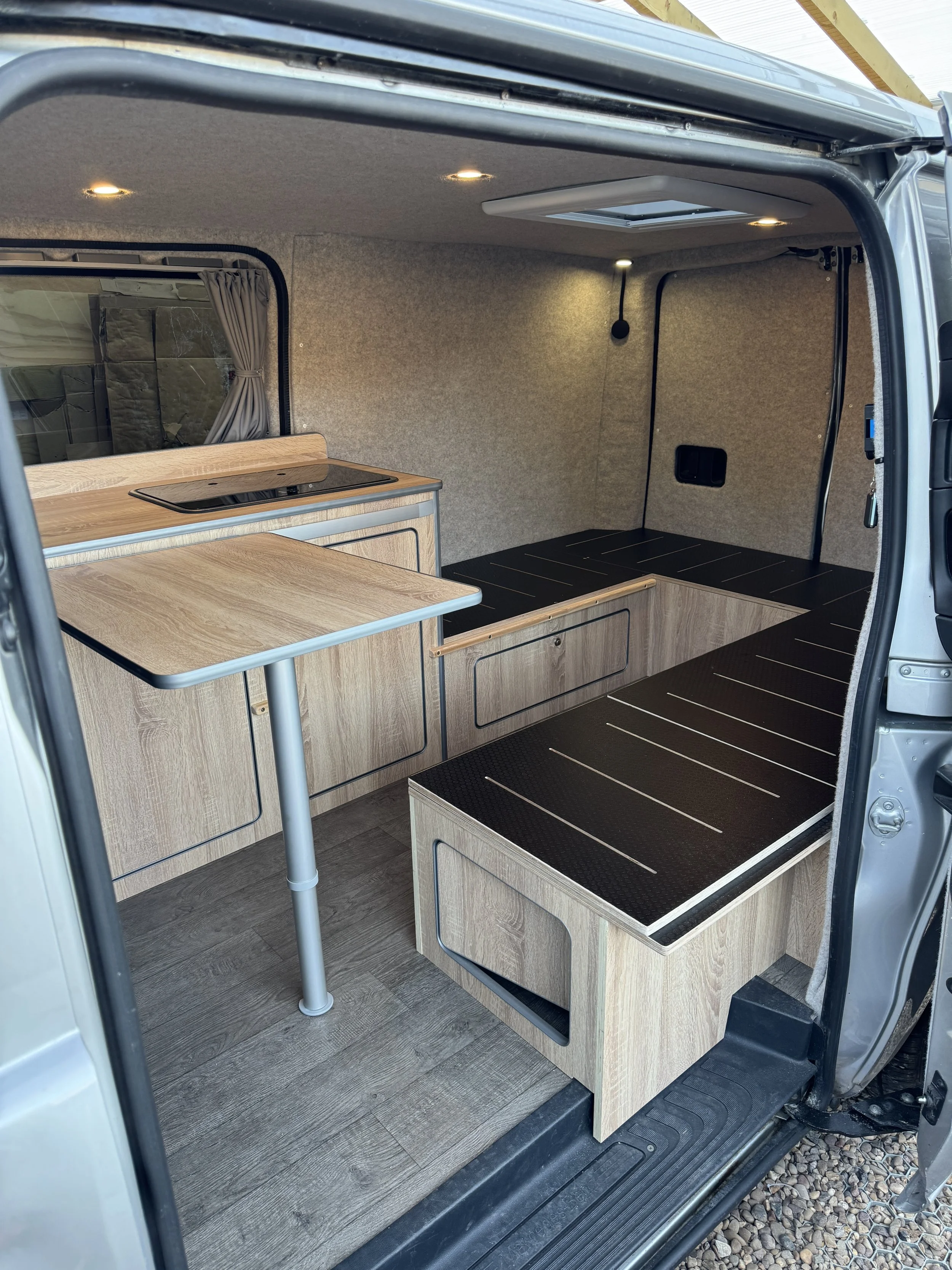 Interior of a compact camper van showing wood-paneled furniture, a small table, a built-in bed platform, and a window with curtains.