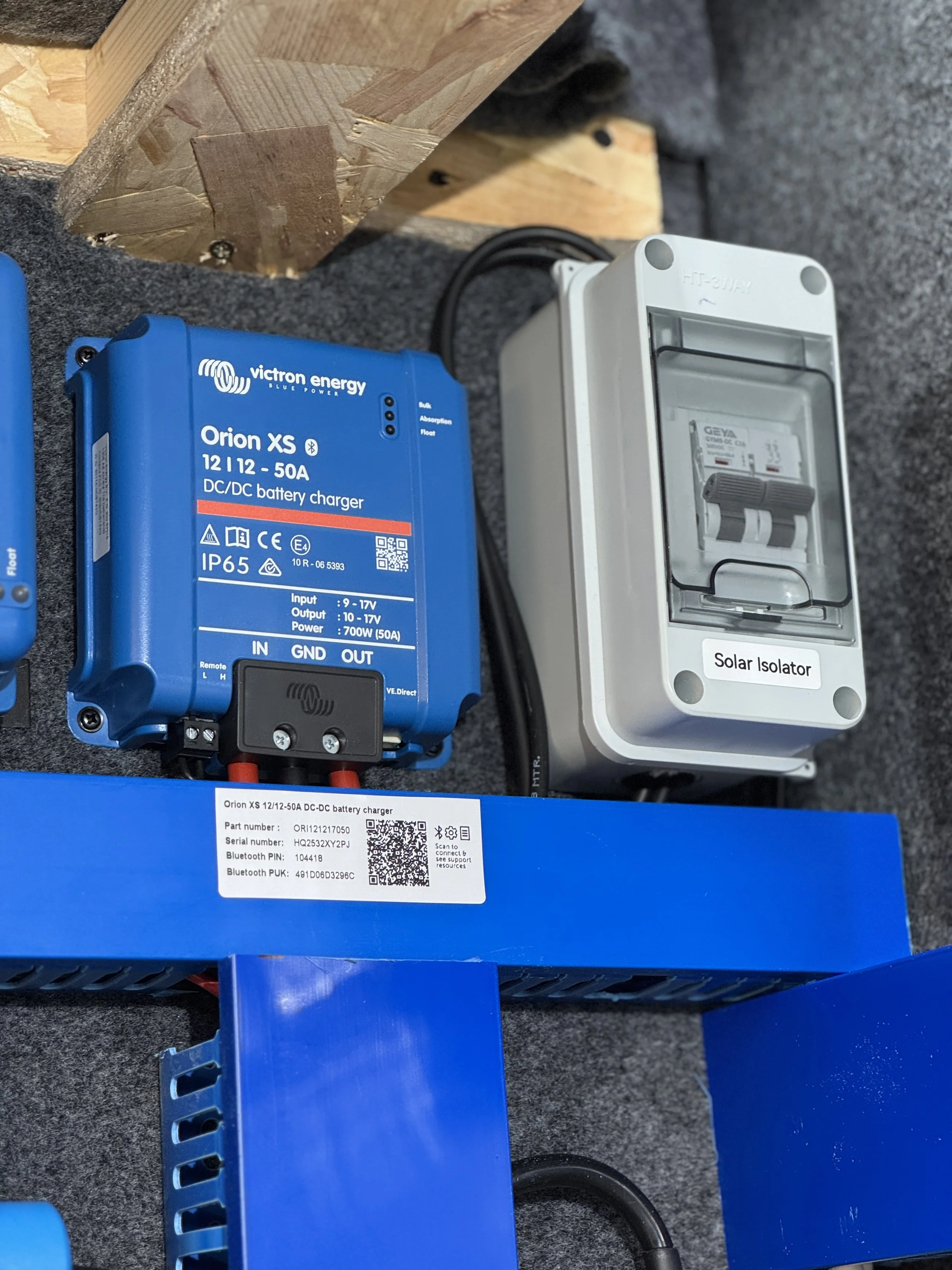Close-up of a blue Victron energy Orion XS DC/DC battery charger and an electrical panel labeled 'Solar Isolator,' mounted on a grey surface with wooden framing above.