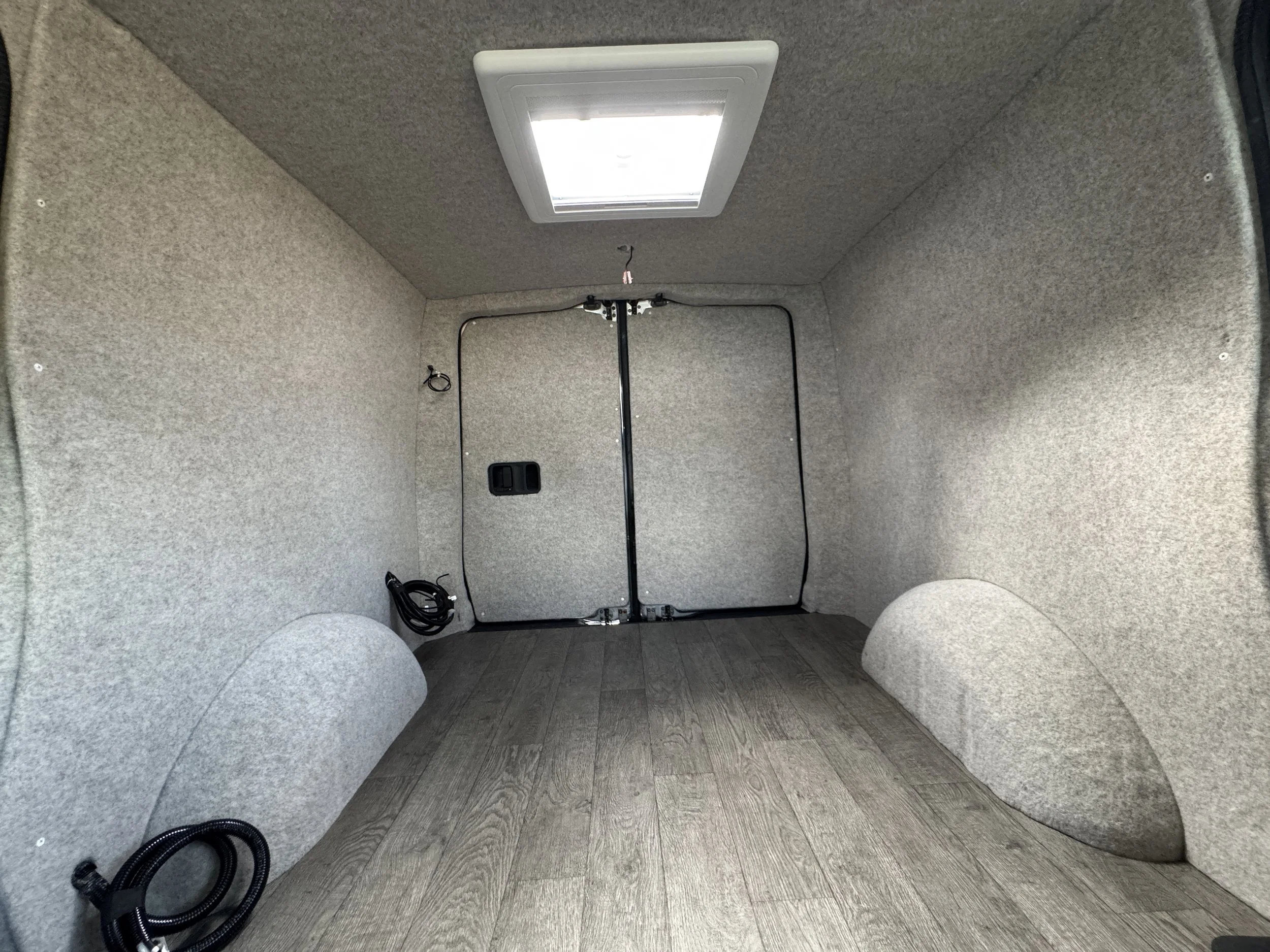 Empty commercial cargo van interior with gray walls and ceiling, wood-look floor, and a roof vent.