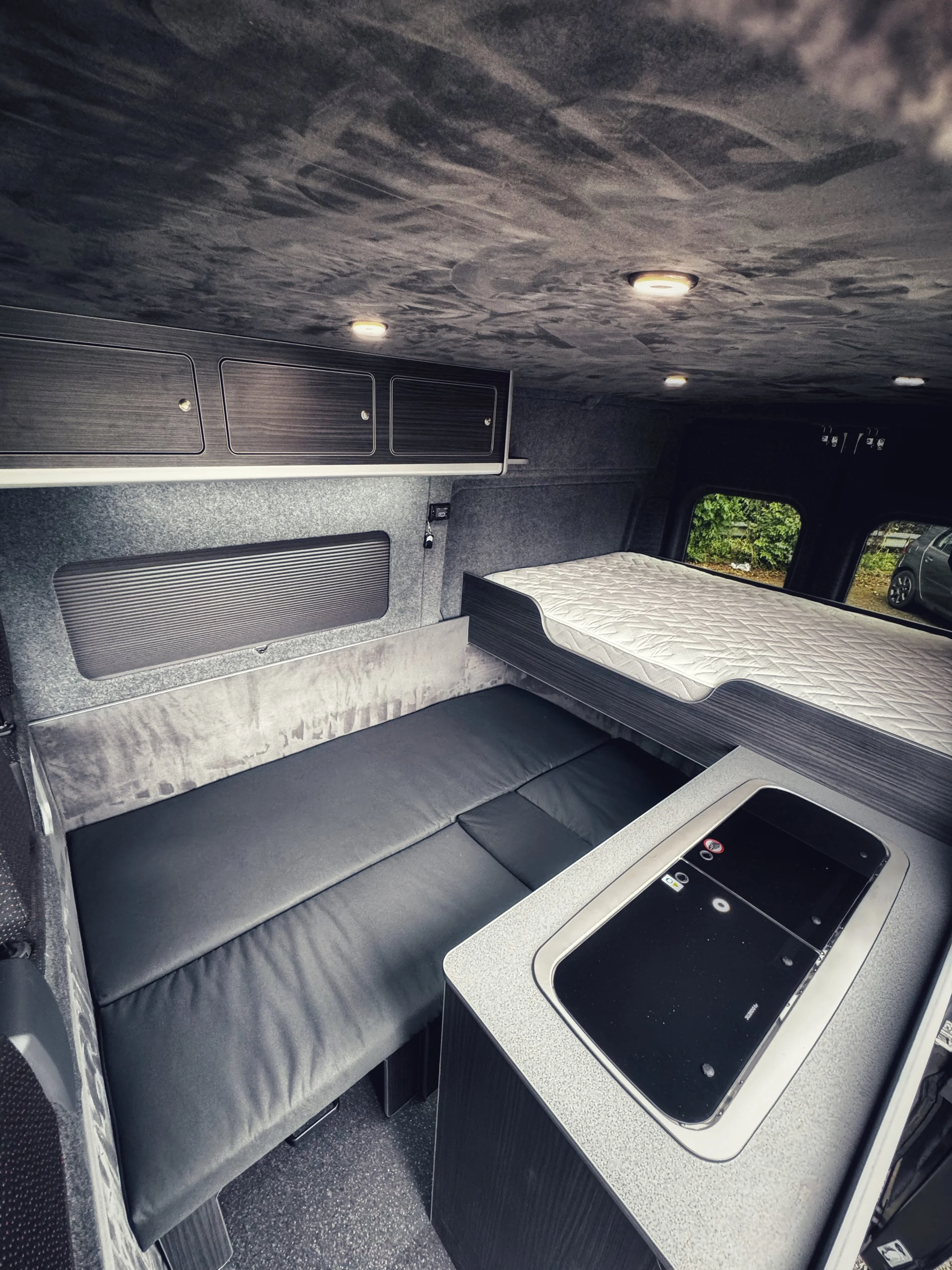 Interior of a van converted into a camper with a sleeping area, overhead cabinets, and a kitchenette.