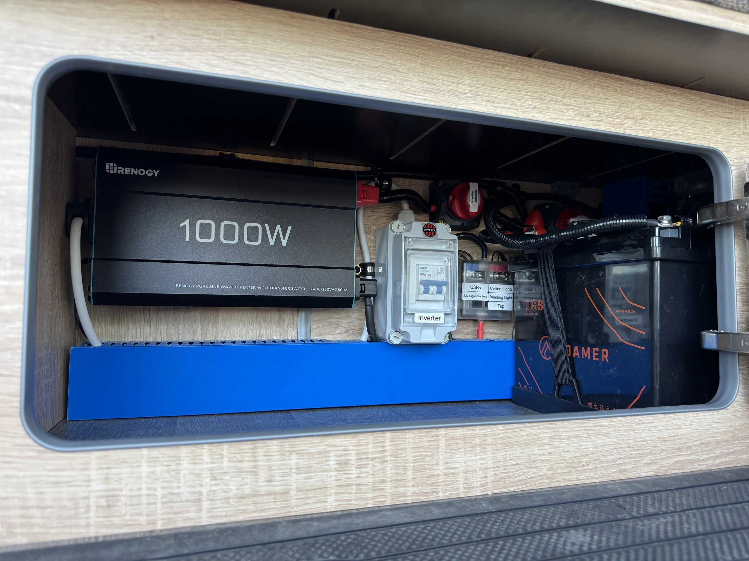 Entertainment system or storage compartment with electronic components, including a 1000W inverter, a blue power strip, a circuit breaker, and various wires and connectors.