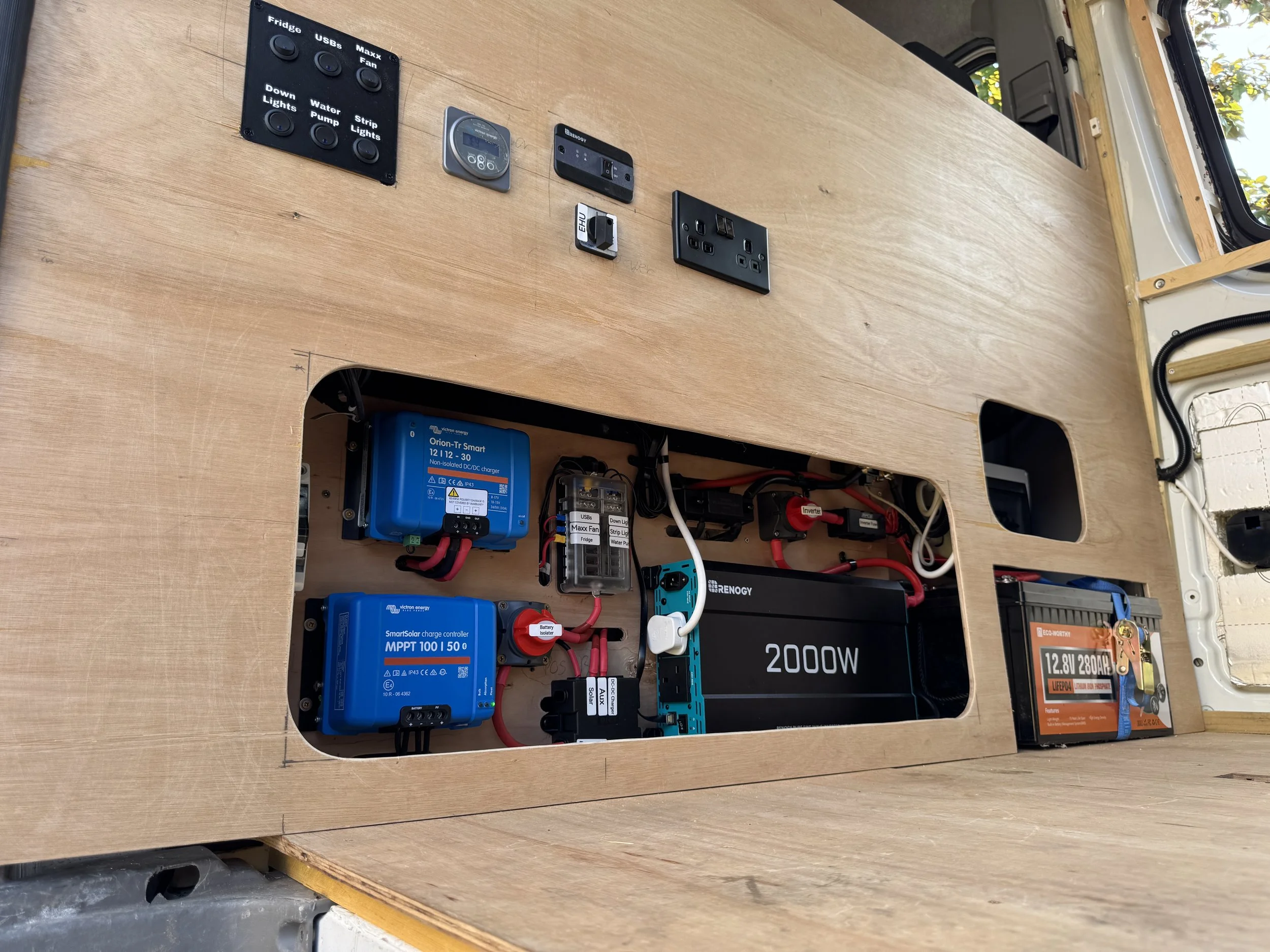 Interior view of a van with an organized electrical system, including a blue inverter, a black 2000W power unit, a battery, and various switches and outlets mounted on a wooden panel.