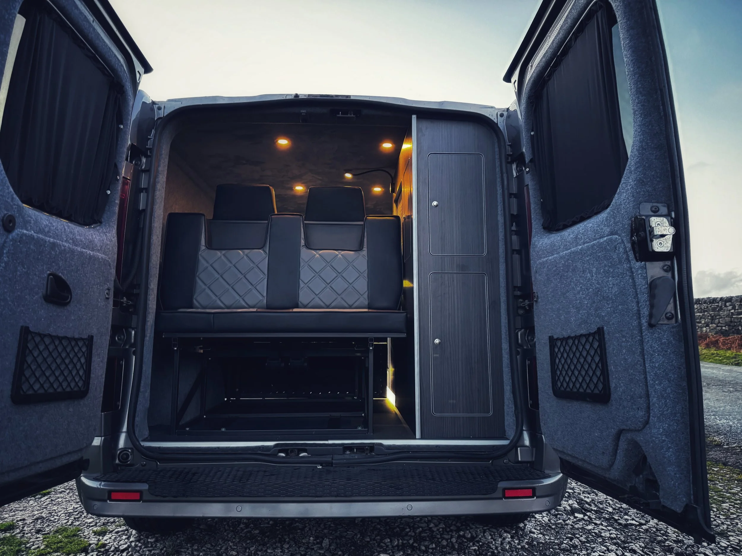 Interior of a camper van with open rear doors, showing seating, storage cabinets, and overhead lighting.