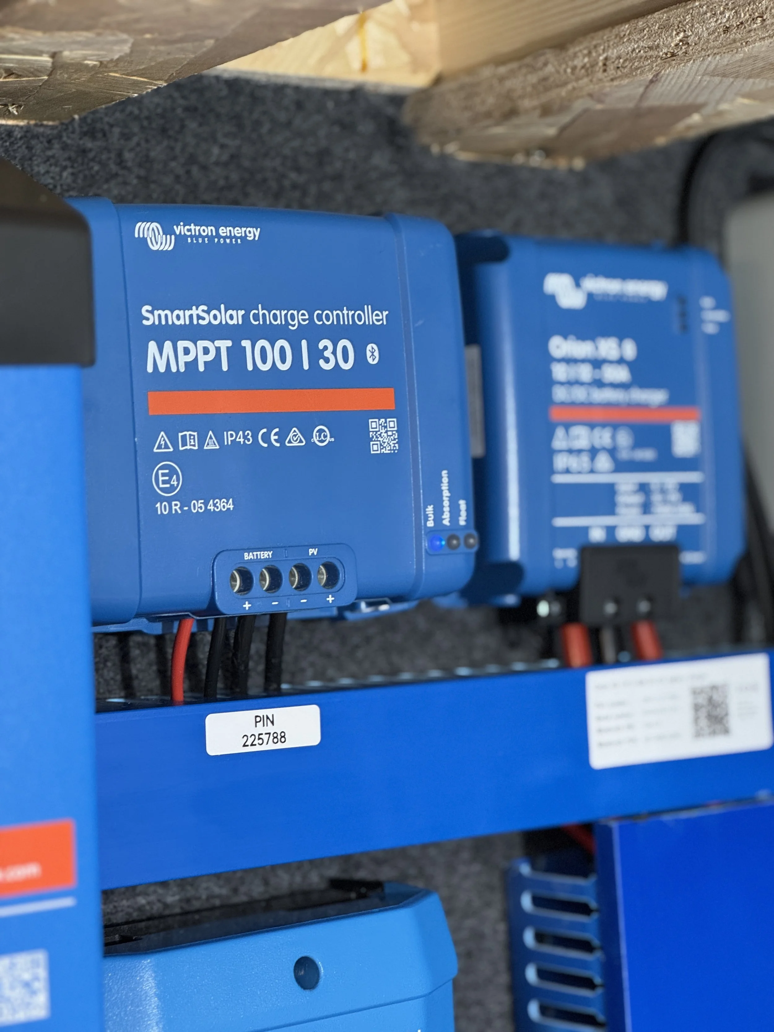 Close-up of blue solar charge controllers mounted on a wall, labeled 'SmartSolar charge controller MPPT 100|30' with various certification symbols, wiring connections, and a PIN sticker.