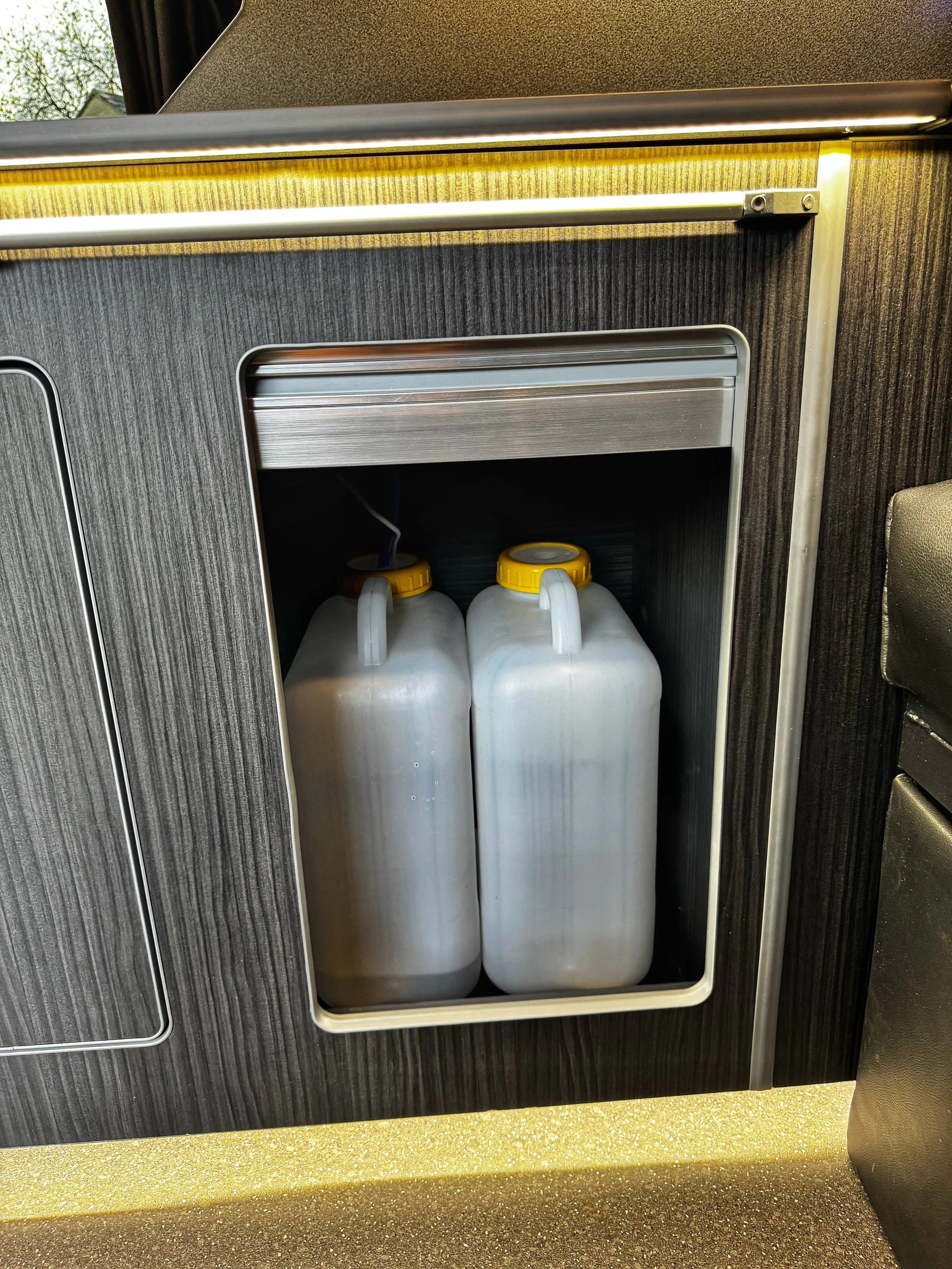 Two white plastic jugs with yellow caps stored in a custom cabinet or compartment, featuring modern wood and metal paneling.