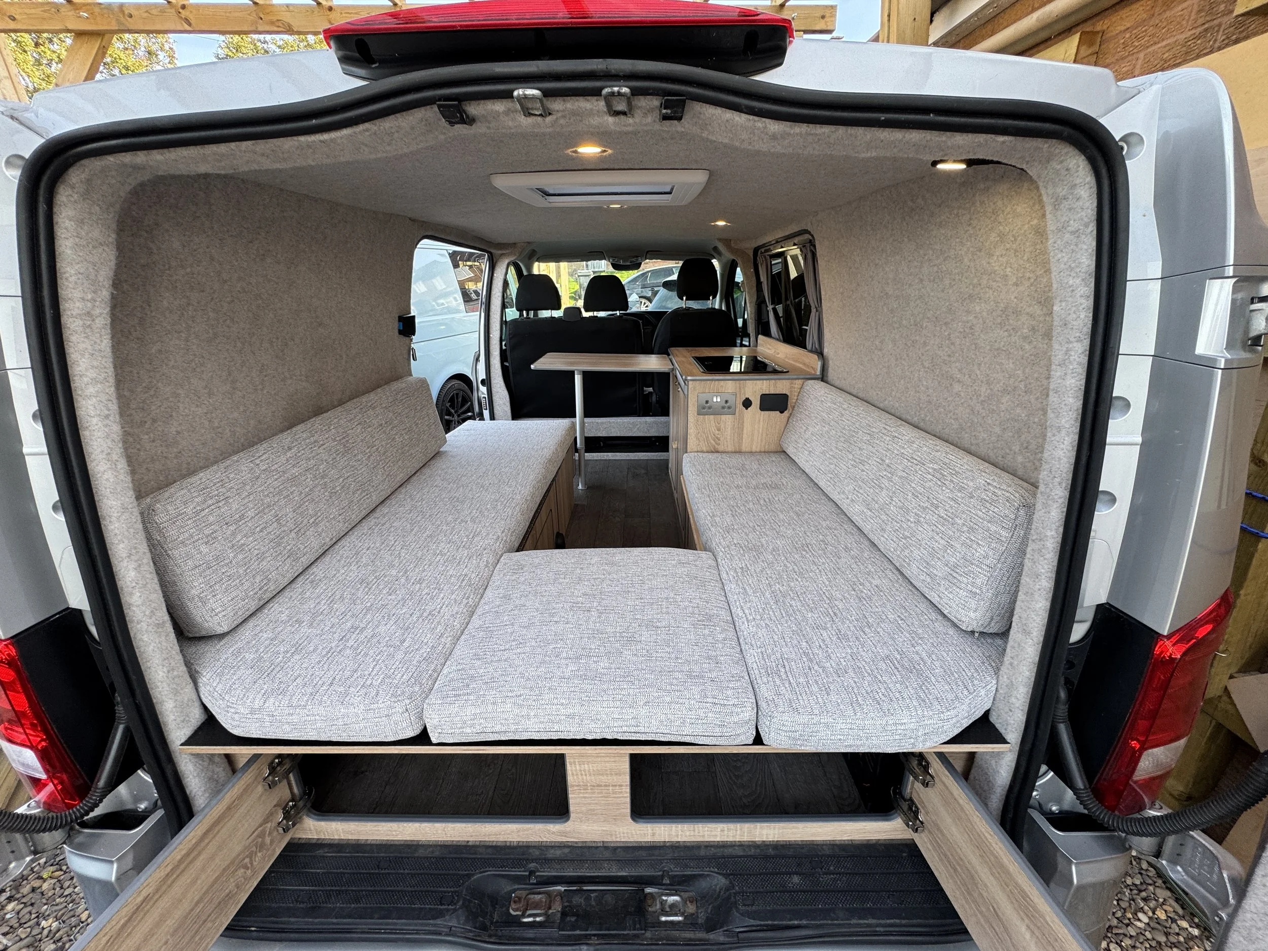 Interior of a custom camper van with seating area, small table, and kitchenette, viewed from the back with the rear doors open.