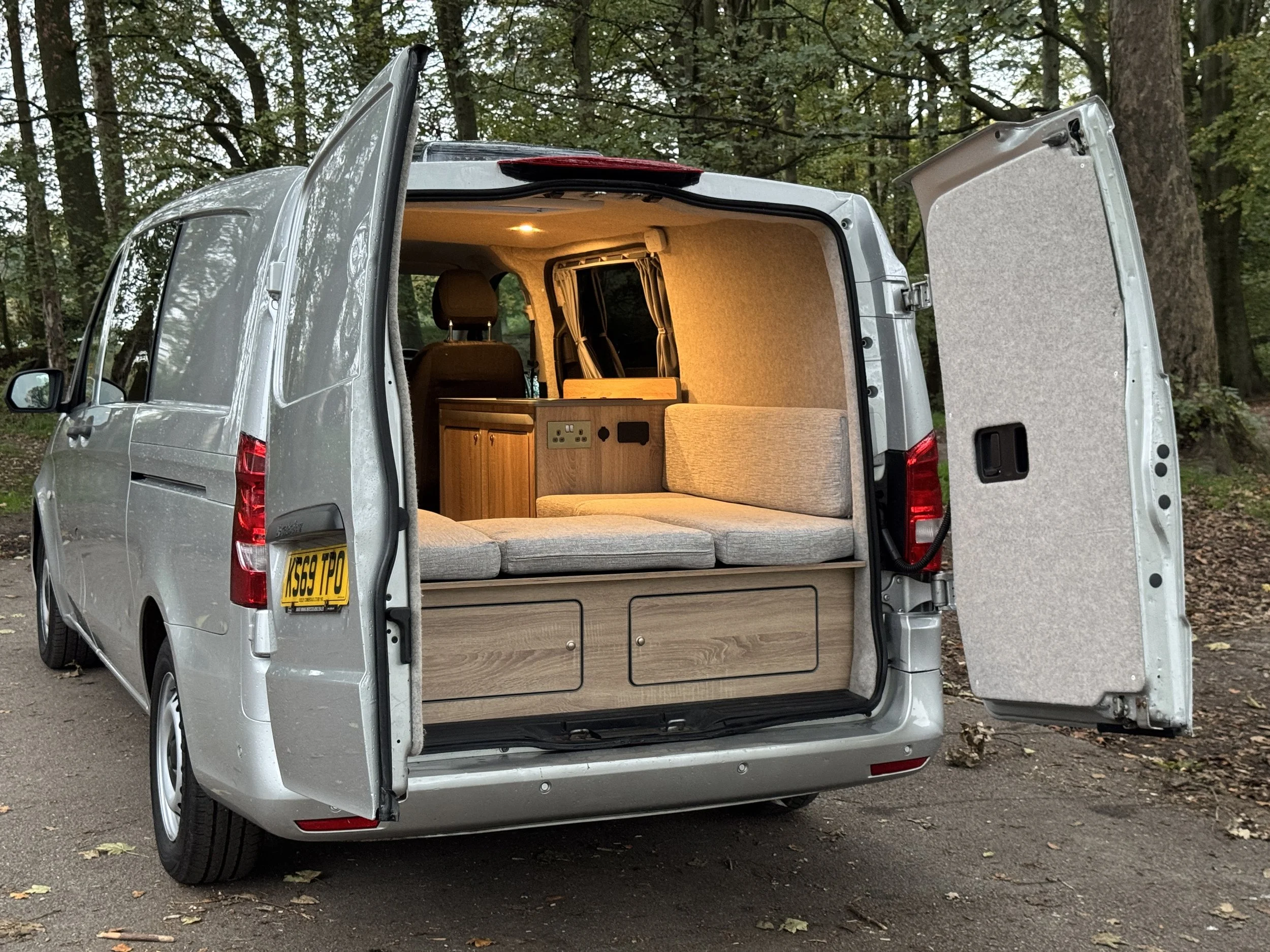Silver camper van with open rear doors showing a cozy interior with seating, wood cabinets, and a power outlet, parked in a wooded area.