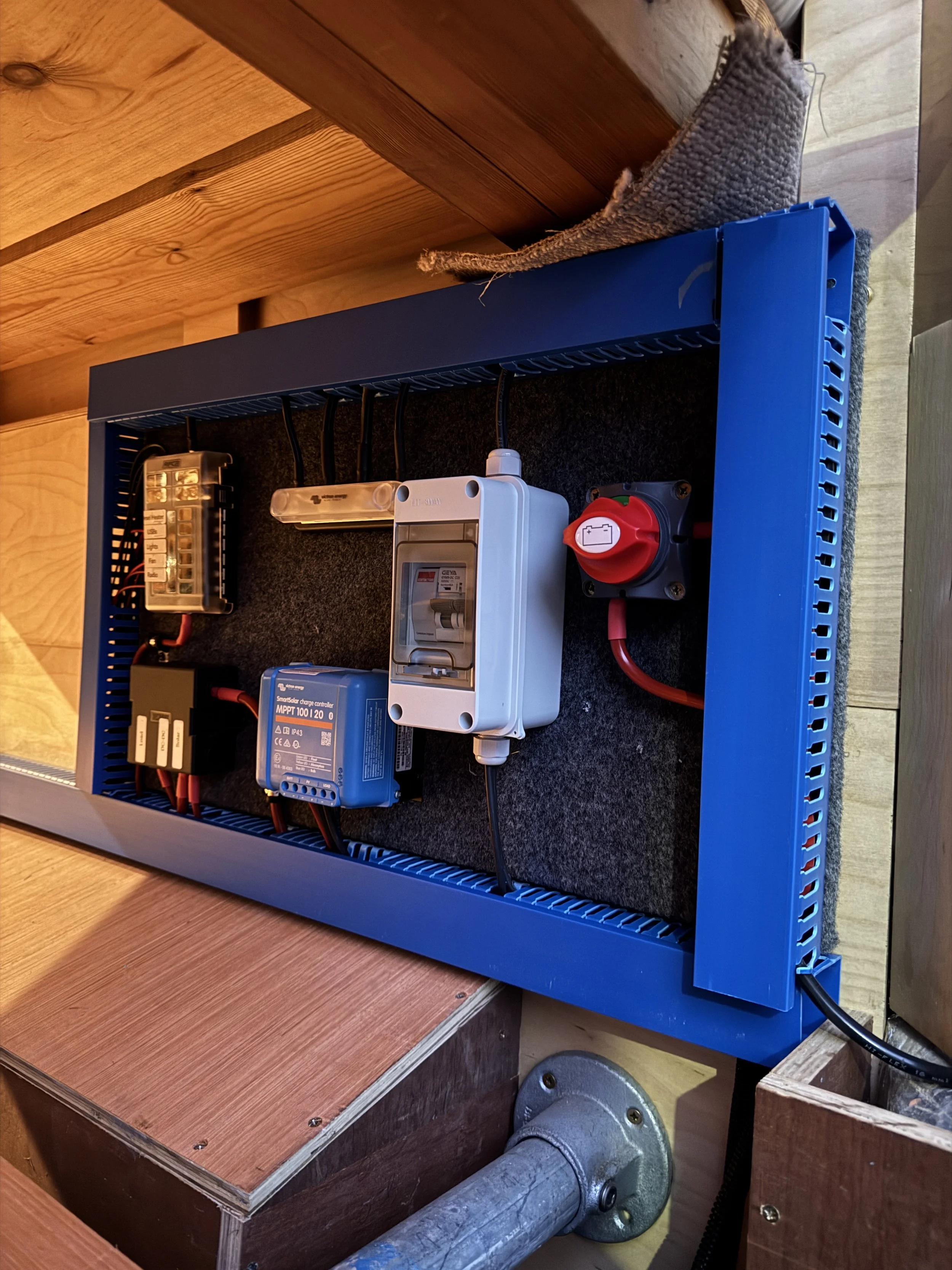 Electrical components mounted inside a blue metal frame on a wooden surface. The components include a circuit breaker, a blue charge controller, a power outlet with a red switch, and various wires. The setup appears to be part of an electrical or sol