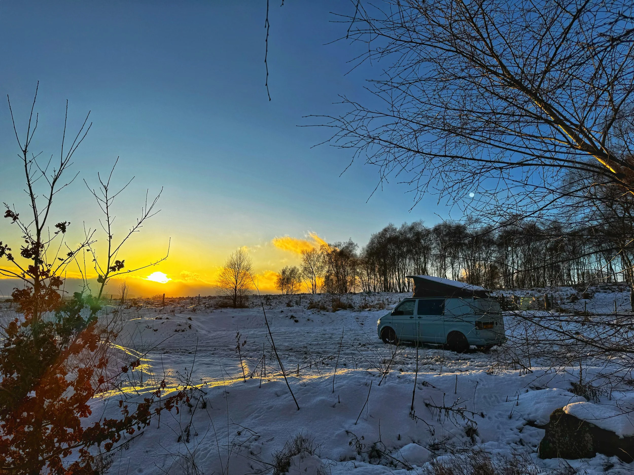 Camper van parked on snowy landscape during sunset with bare trees and clear sky.