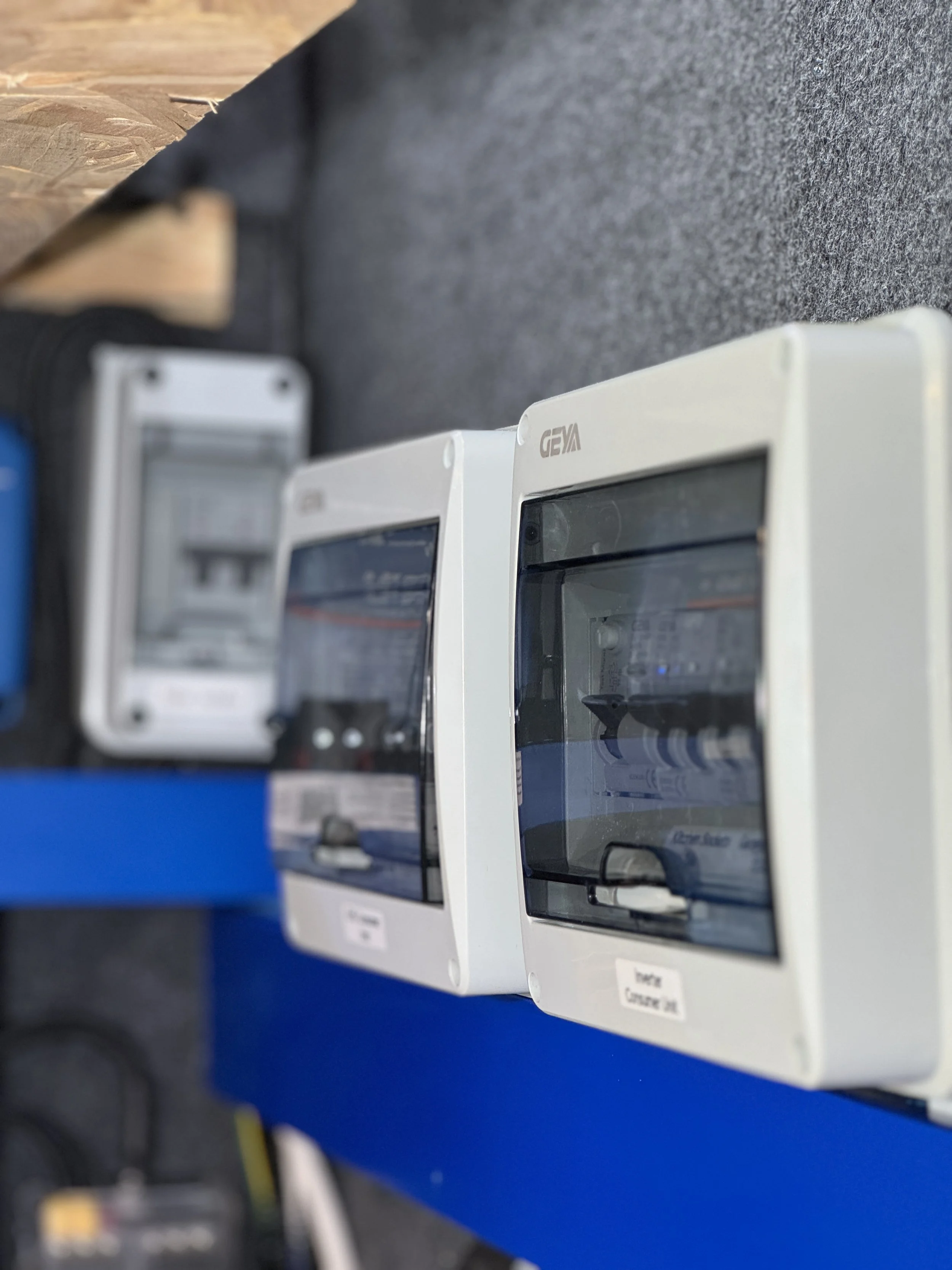Multiple electrical meters with transparent covers mounted on a blue panel, placed on a gray carpeted floor.