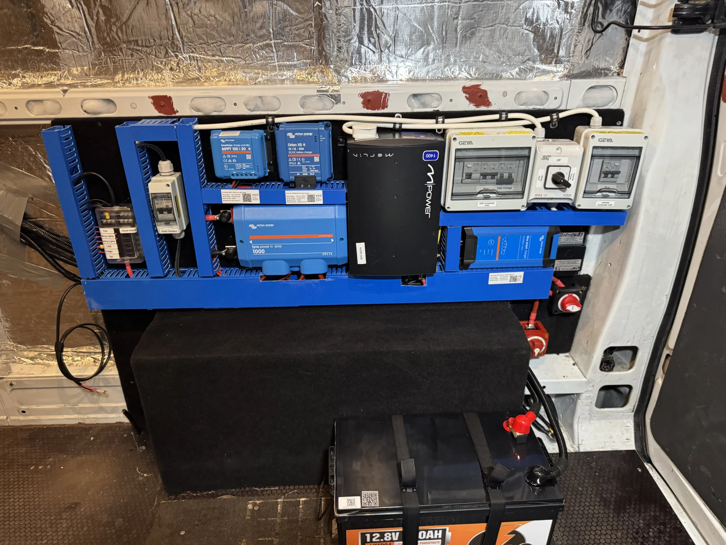 Electrical control panel with various blue and white components, circuit breakers, and wiring, mounted on a metal panel with a black foam covering at the base and a black battery at the bottom.