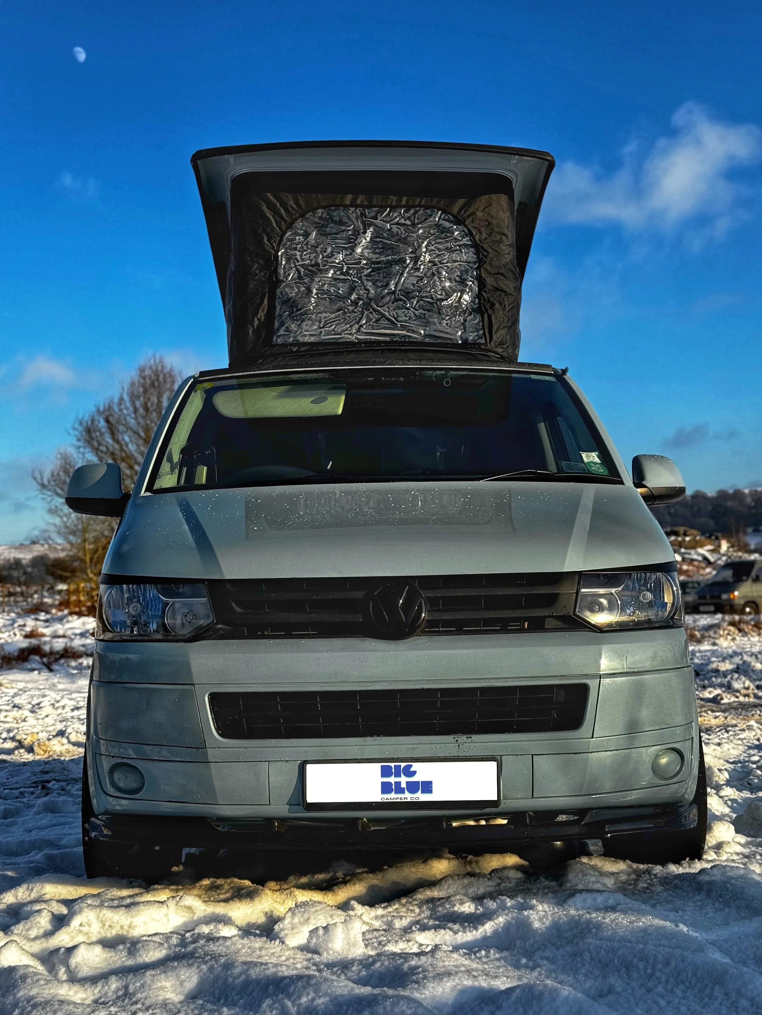 Front view of a gray camper van with its pop-top roof open, parked on snow under a clear blue sky.