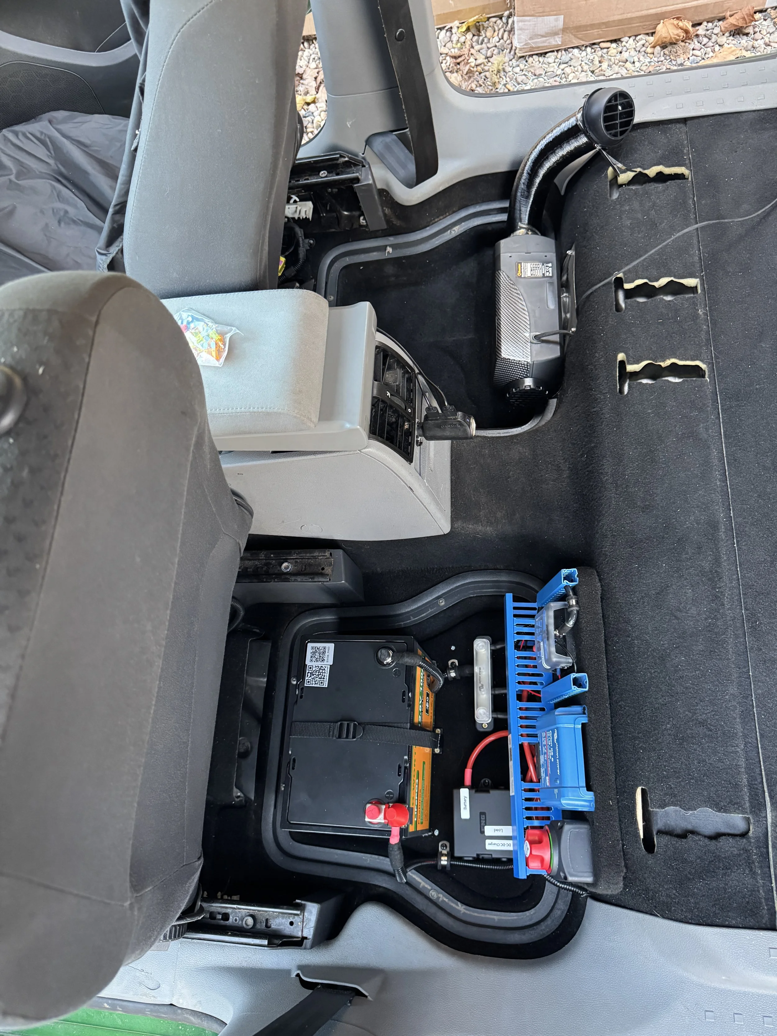 The back of a vehicle interior showing a large battery pack with wiring, a blue connector, and a power inverter or charger system installed on the floor behind the front seats.