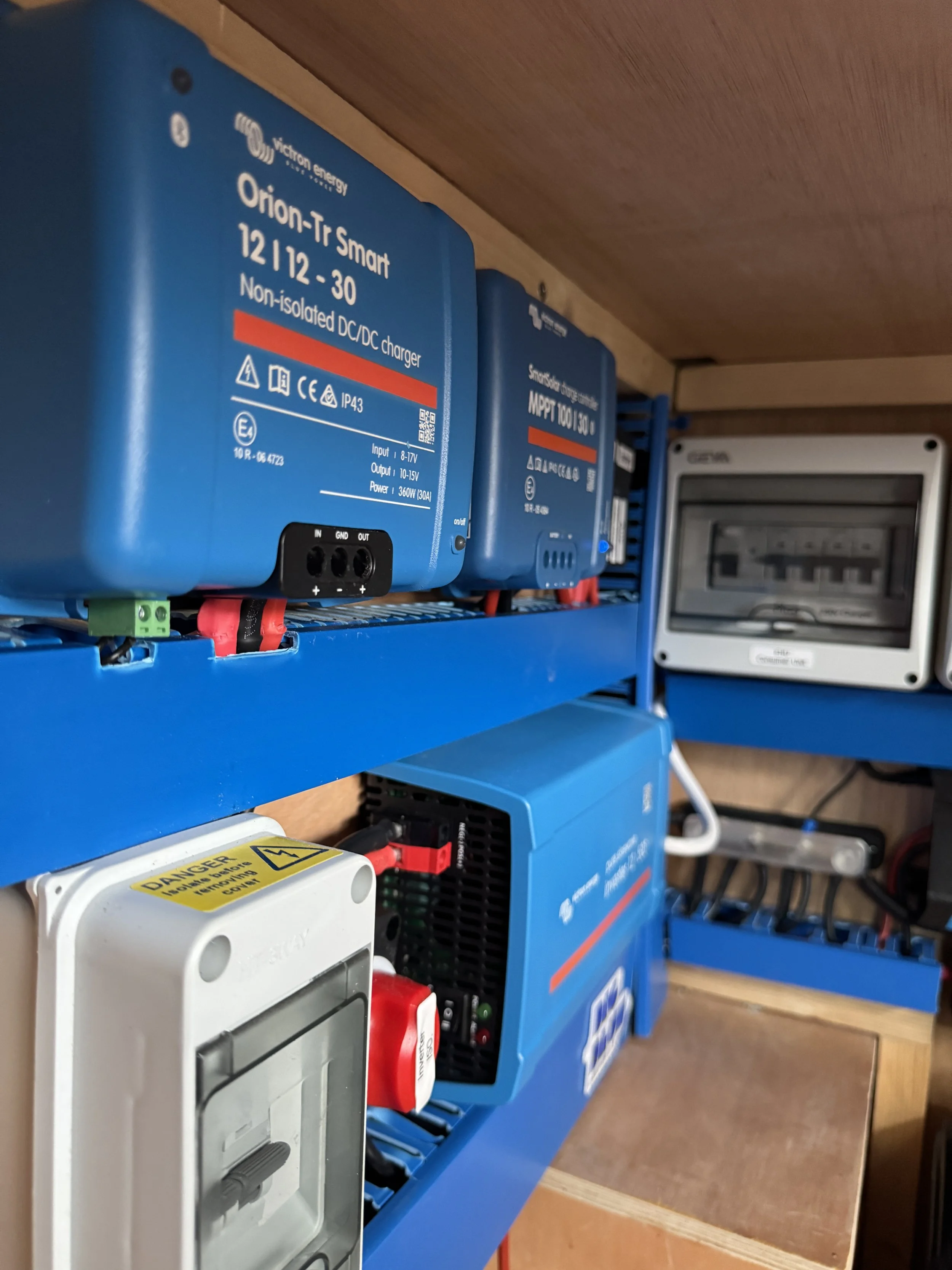 12v 230v Campervan electrical panel with, including a blue Orion-Tr Smart inverter, a white electrical box with a warning label, a blue power supply module, and an electrical panel in the background.