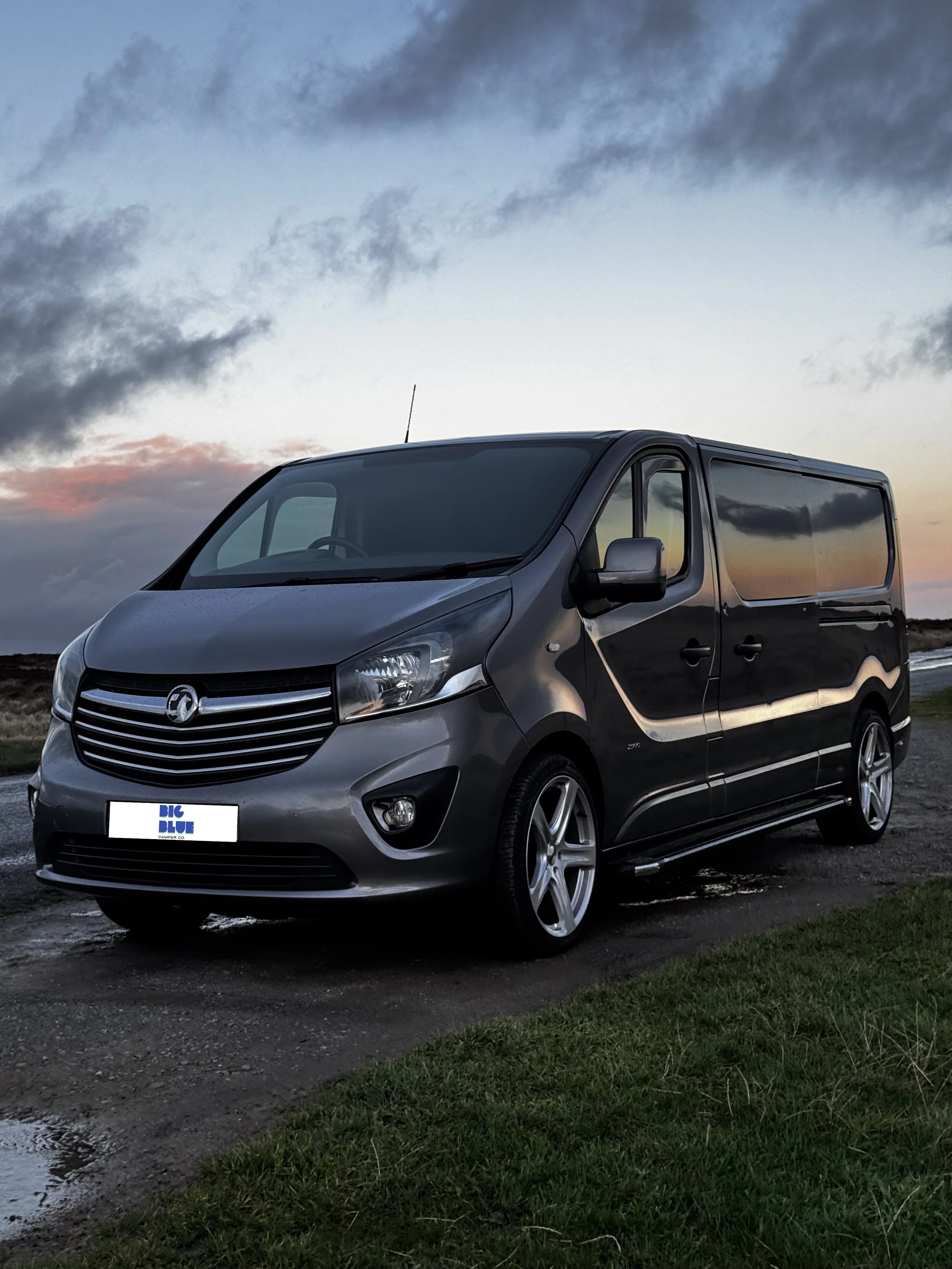 Gray van parked on a countryside road at sunset, with grass in the foreground and a cloudy sky in the background.