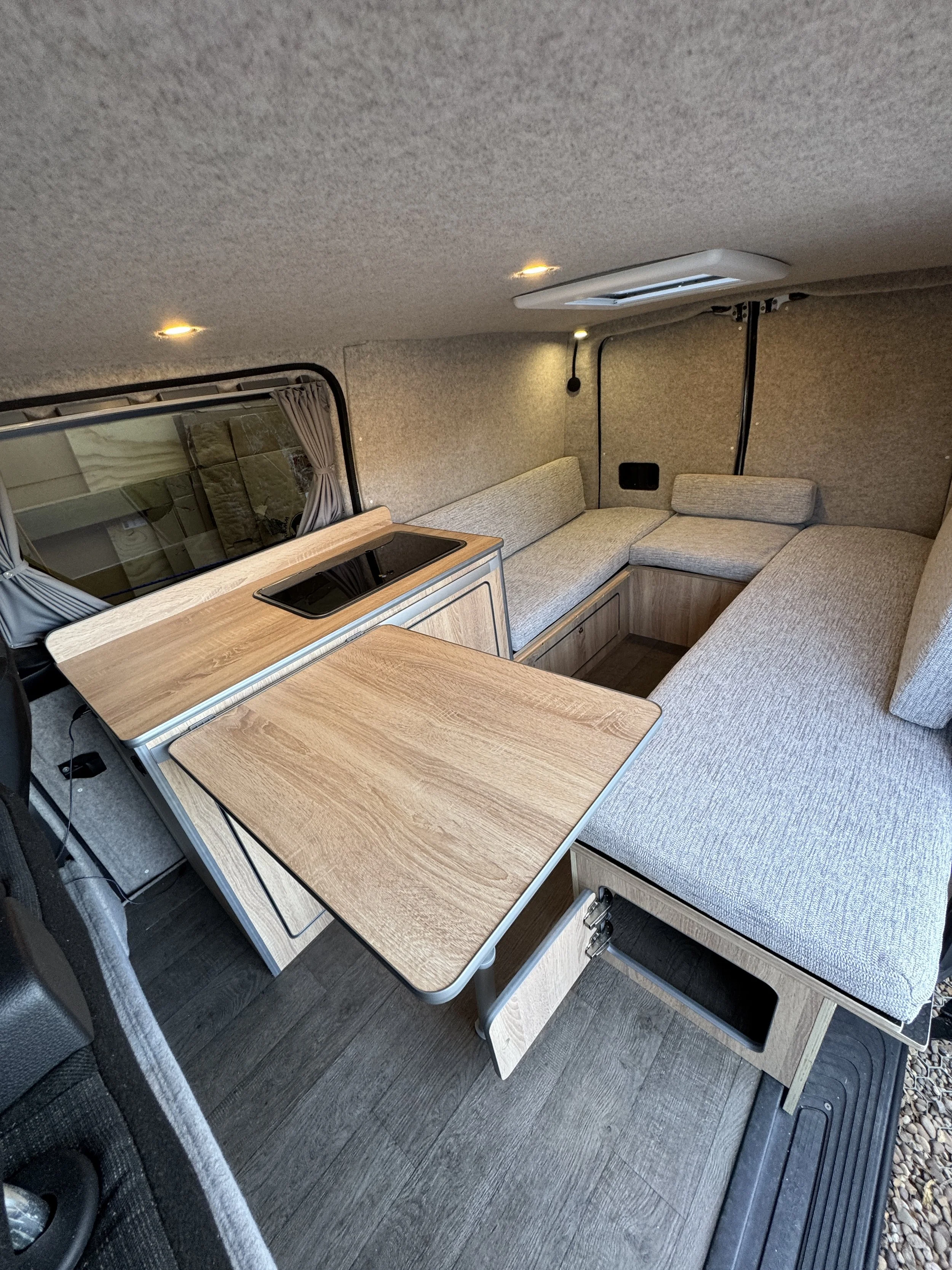 Interior of a compact camper van with a U-shaped sofa, wooden table, small kitchen area, window with curtains, and ceiling lights.