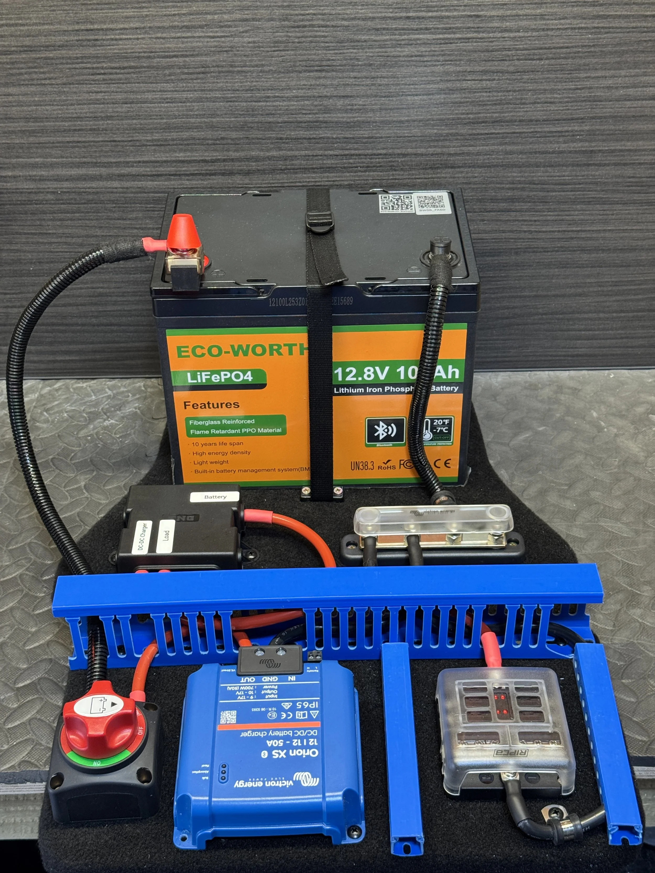 A setup of a battery test system with a large Eco-Worth 12.8V, 10Ah lithium iron phosphate battery connected to a blue variable speed drive, cables, and control devices on a black surface with a gray textured background.