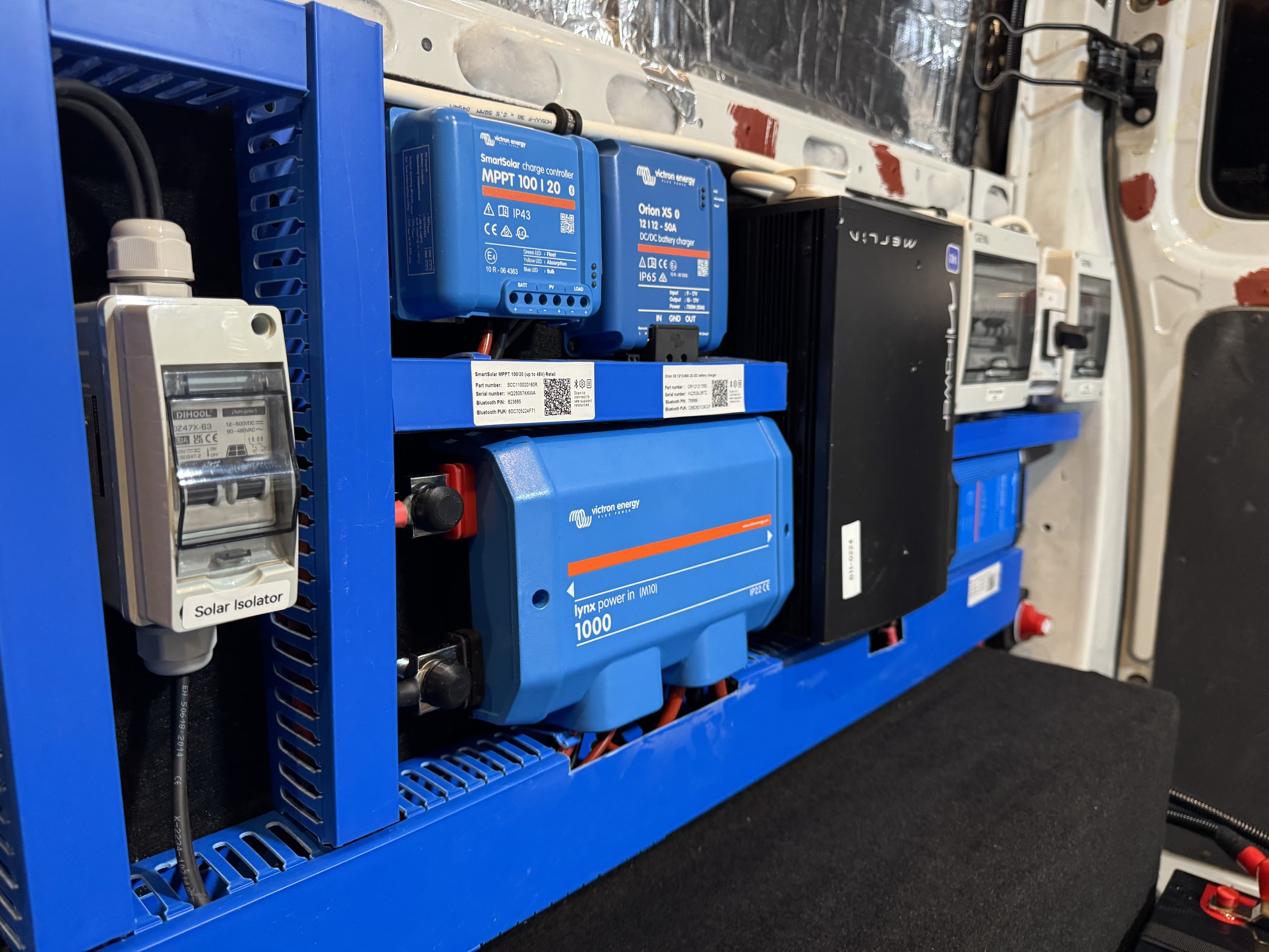 Close-up of electrical components inside a blue metal chassis, including a blue inverter, blue charge controller, black power supply, and a solar isolator switch, mounted on a panel with wires connected to each device.