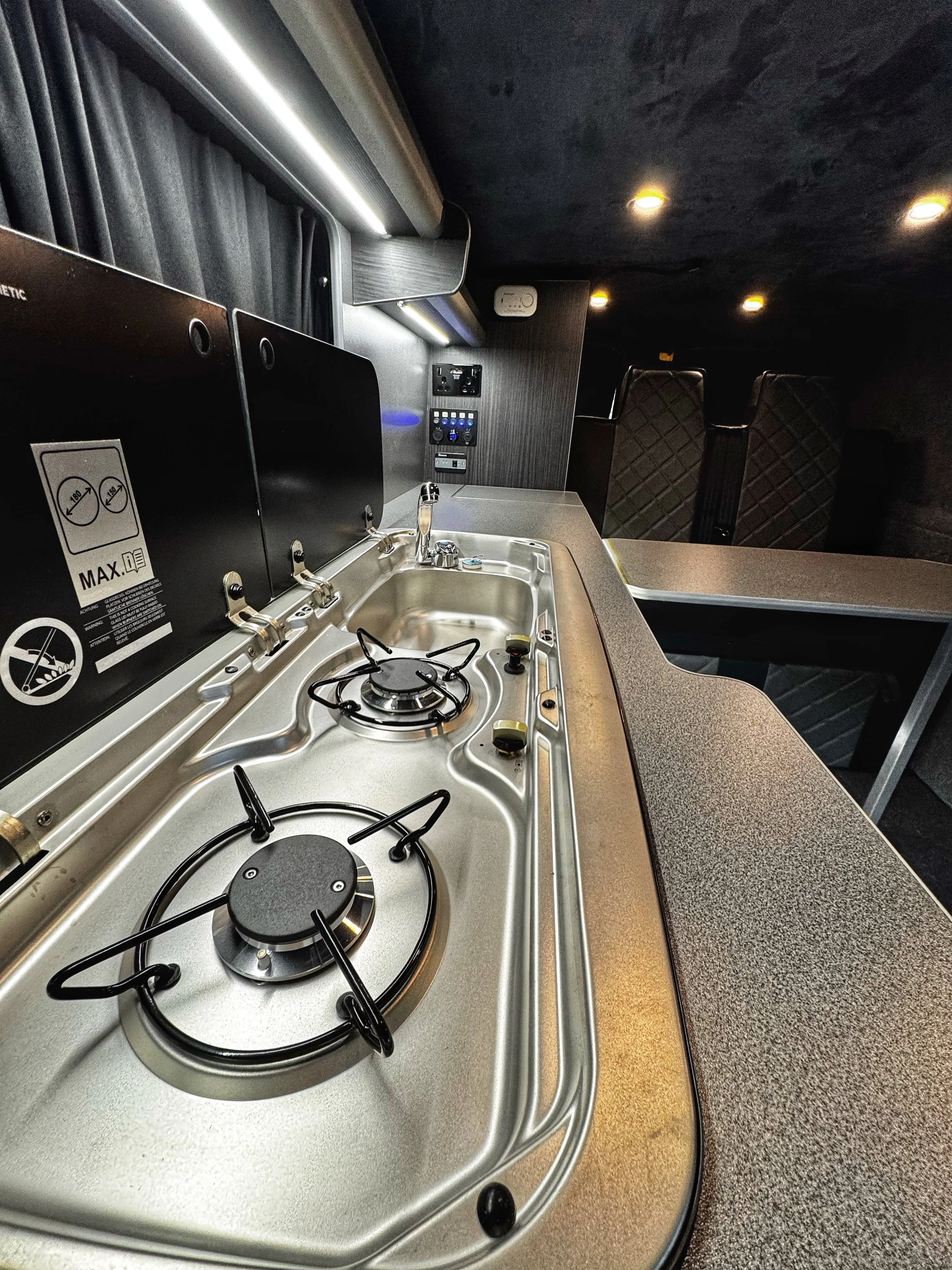 Interior of a camper van featuring a compact kitchen with a two-burner gas stove, a sink, countertop, overhead lighting, and foldable cover panels.