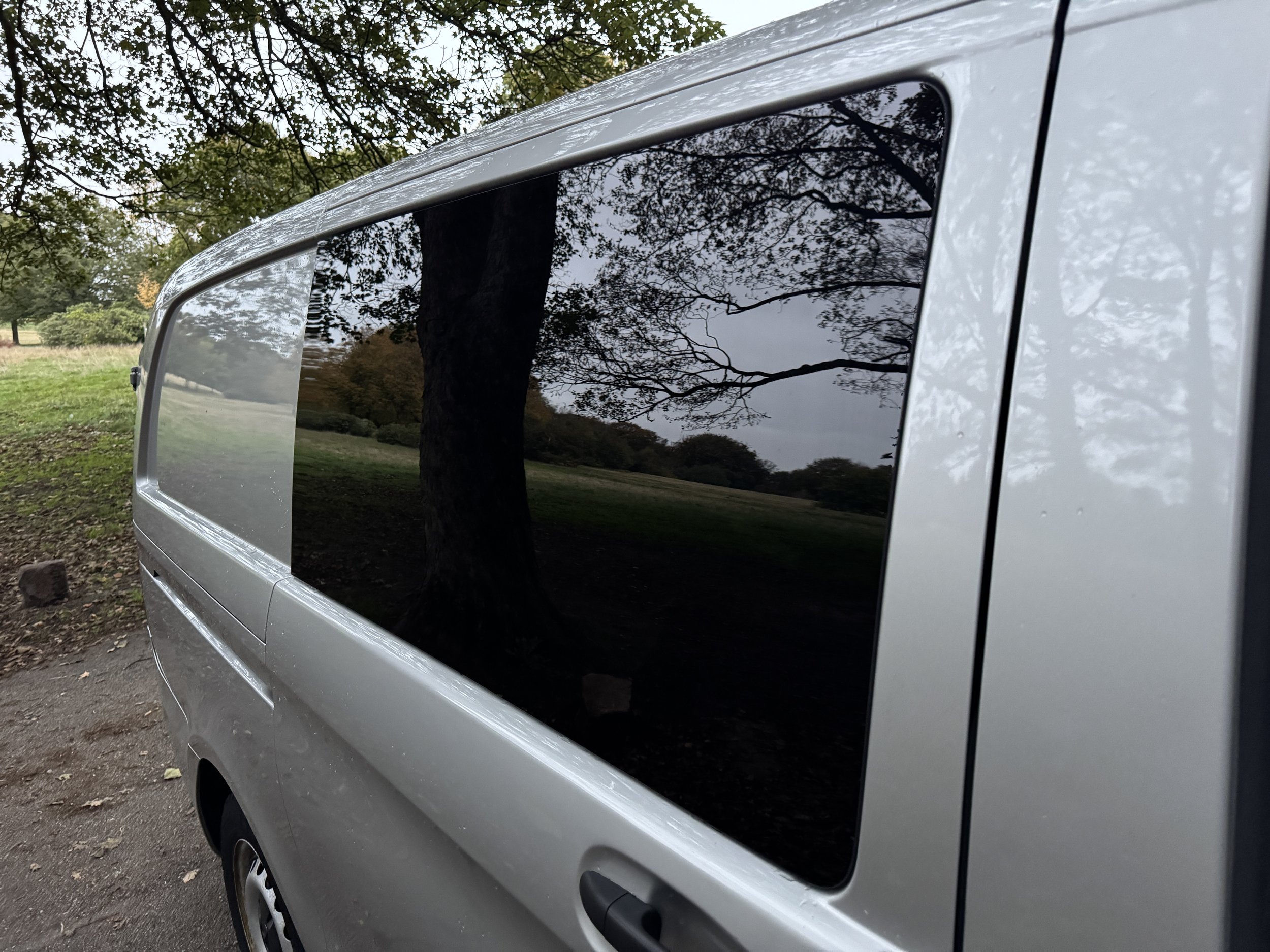 Close-up of a silver van's tinted window reflecting trees and the outdoors, with a grassy area and trees in the background.
