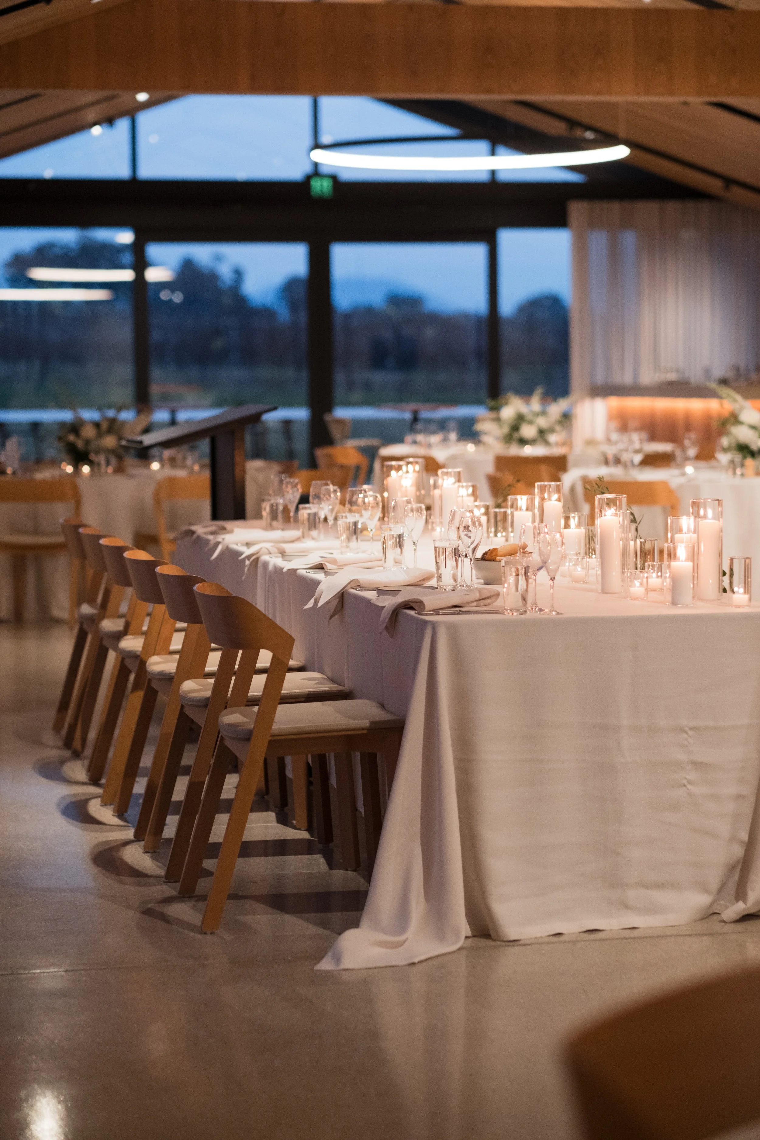 Elegant banquet table set for a formal event with white tablecloth, candles, wine glasses, and floral centerpieces, inside a modern venue with large windows.