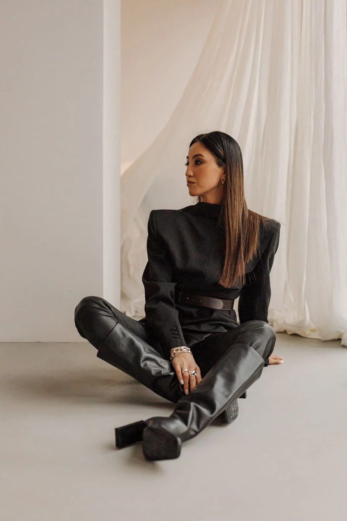 A woman wearing a black blazer and leather pants sitting on the floor in front of a white wall and curtains.