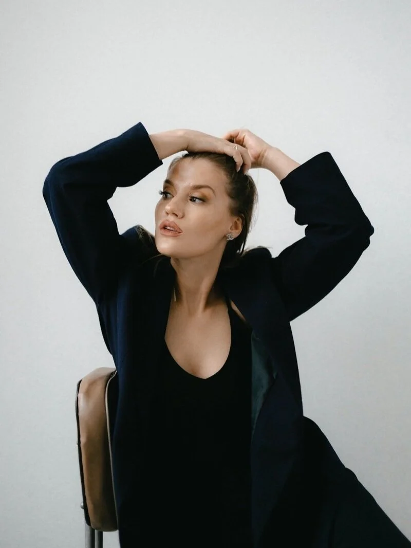 A woman sitting on a chair with her right arm raised, touching her head, wearing a black top and blazer, with a plain white background.