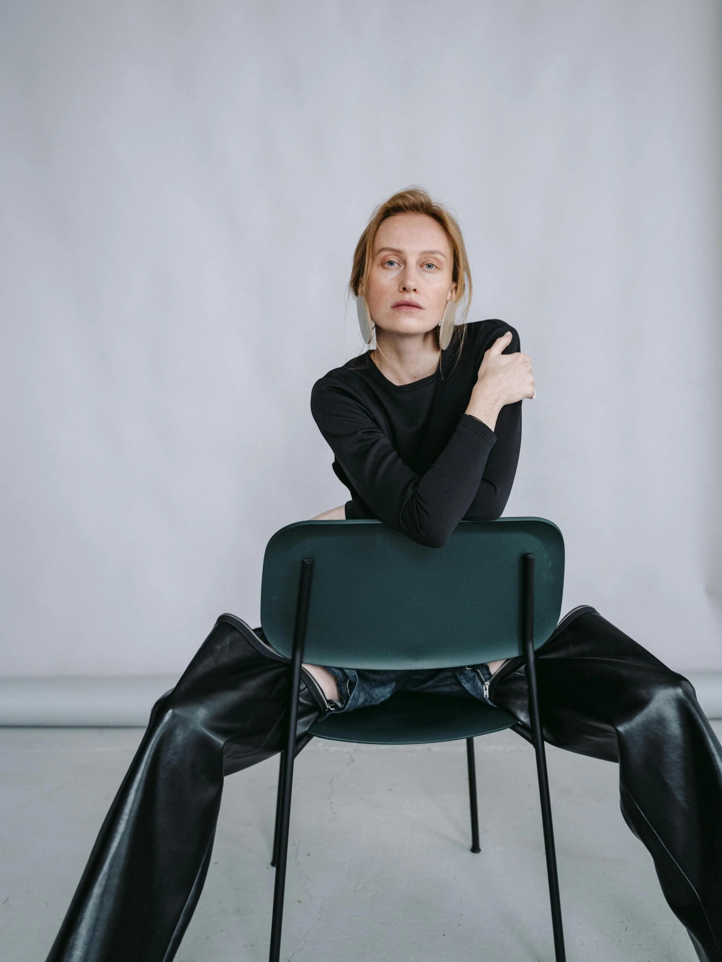 A woman with light skin and red hair, dressed in black top and leather pants, sitting backwards on a green chair, looking directly at the camera with a neutral expression, arms crossed and one hand on her shoulder, against a plain light grey wall background.