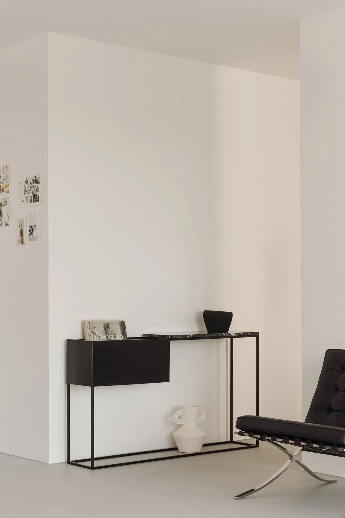Minimalist interior with a black console table, white vase, black chair, and small photo collage on white wall.