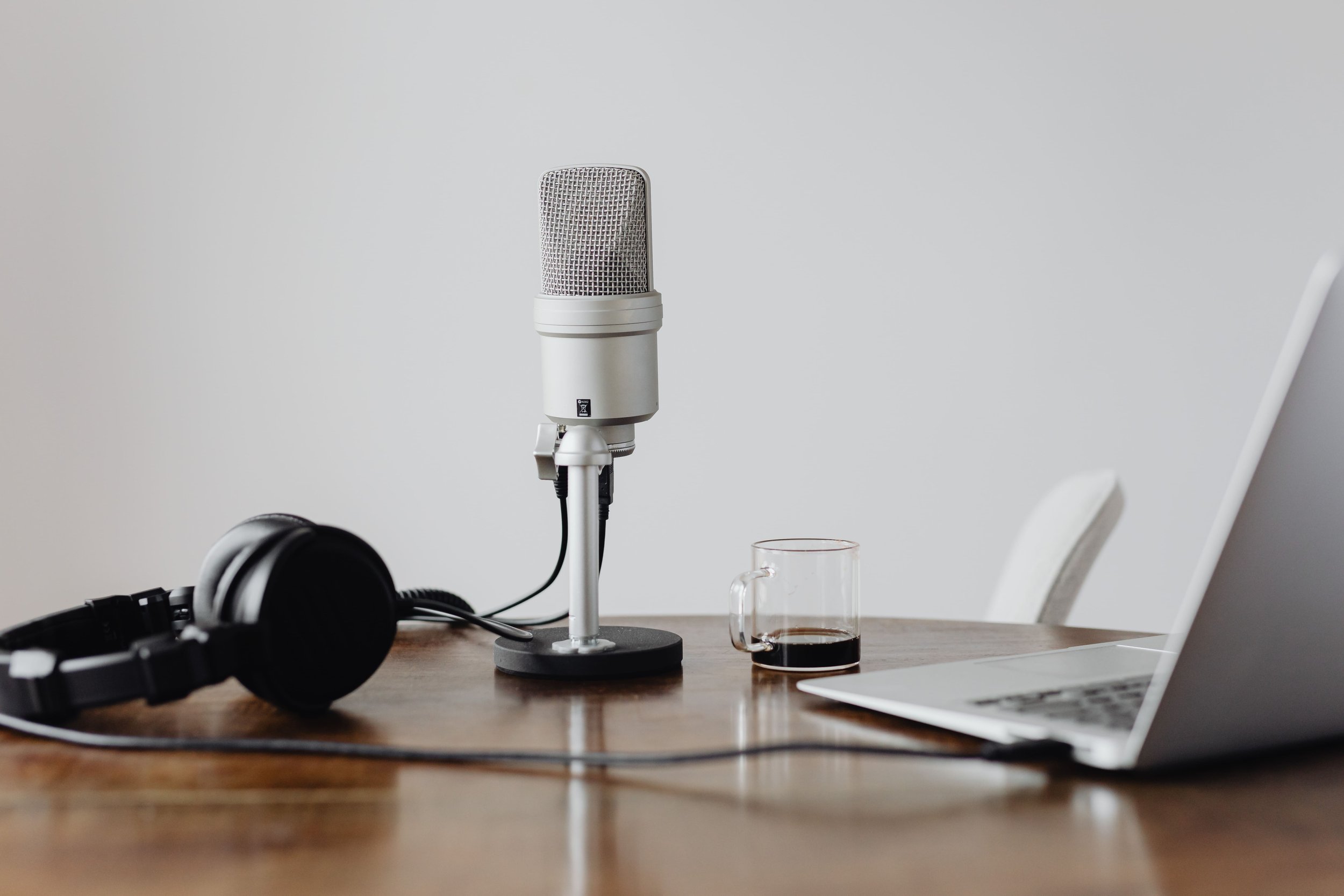 A podcast setup on a wooden table with a microphone on a stand, a pair of headphones, an open laptop, a clear glass mug with coffee, all against a plain white background.
