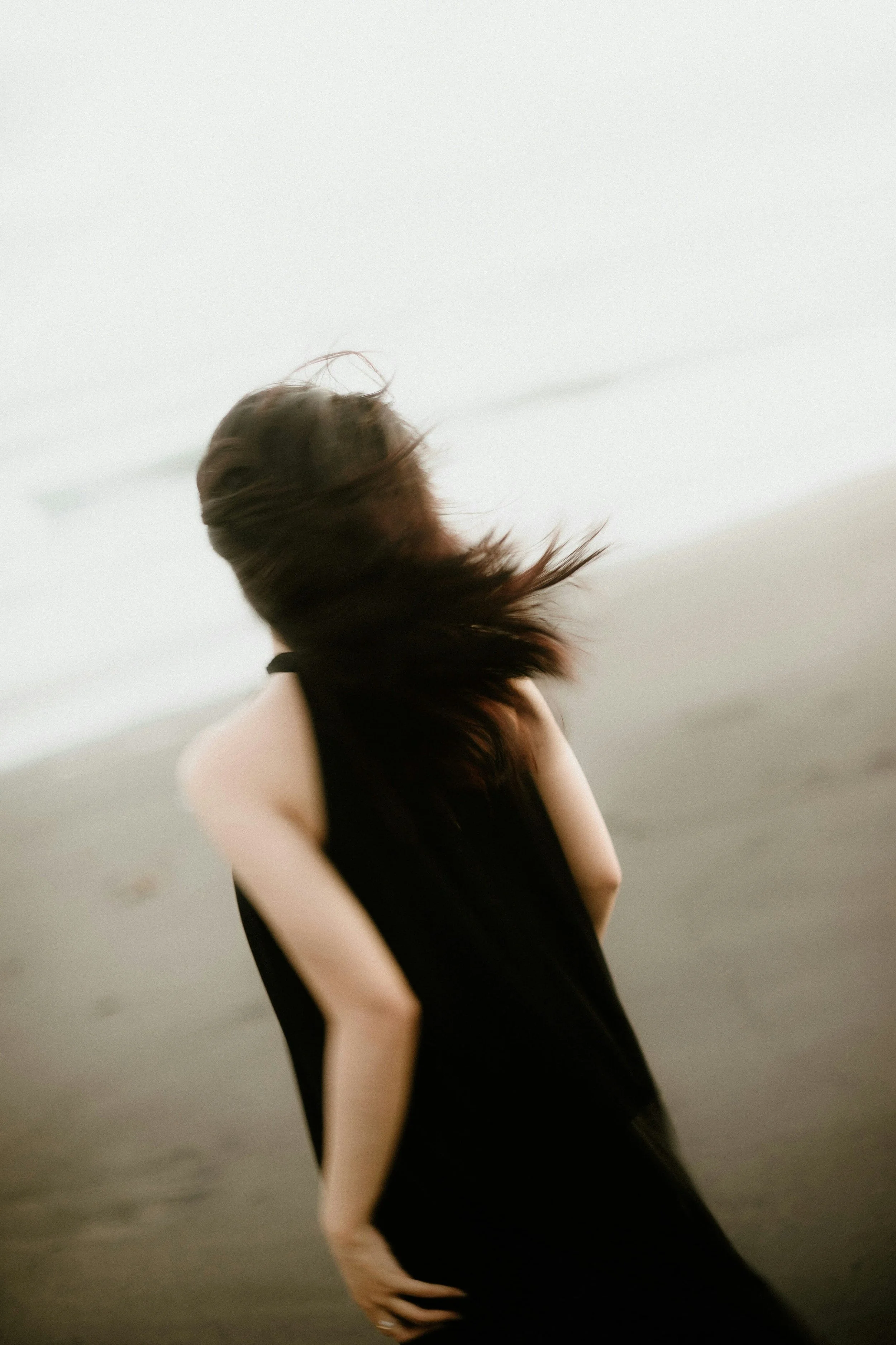 A woman with long, dark hair is standing on a beach, facing away from the camera, with her hair blowing in the wind.