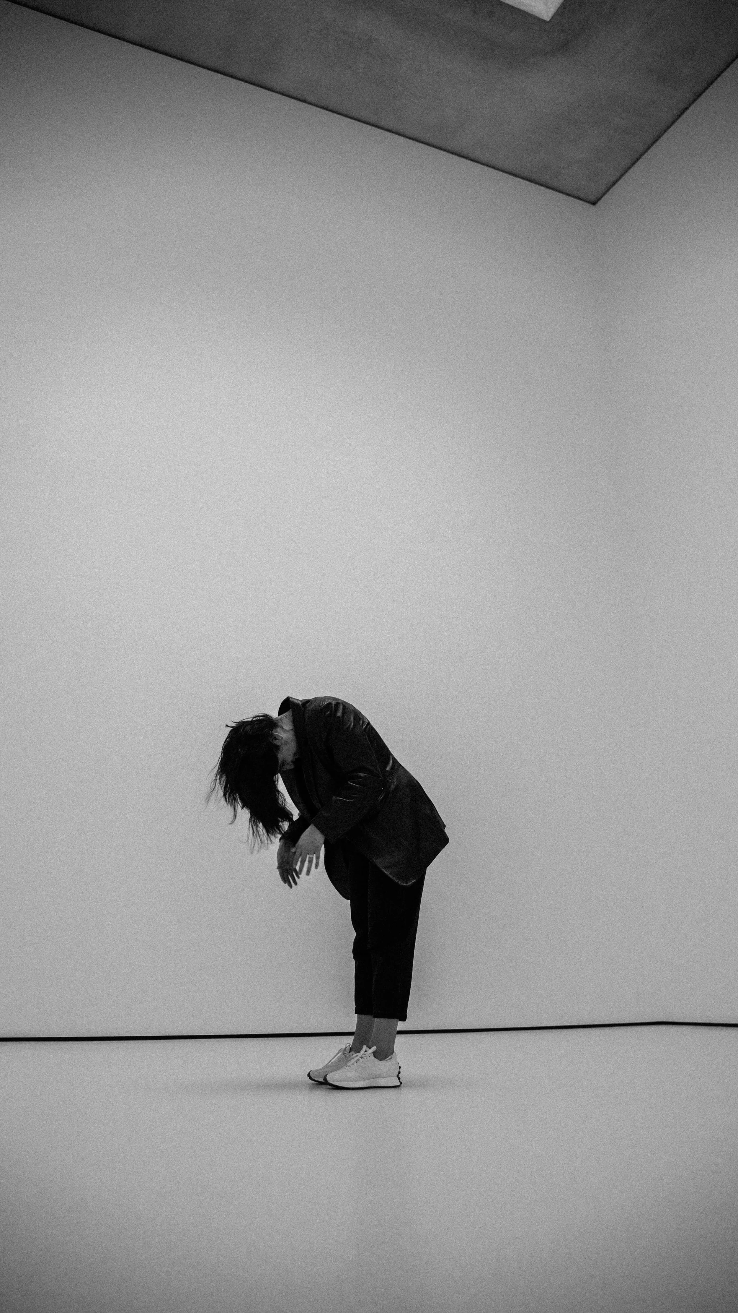 A person with long hair dressed in a black jacket, black pants, and white sneakers, leaning forward with arms crossed and head bowed in a minimalist, empty room with white walls and a dark ceiling.