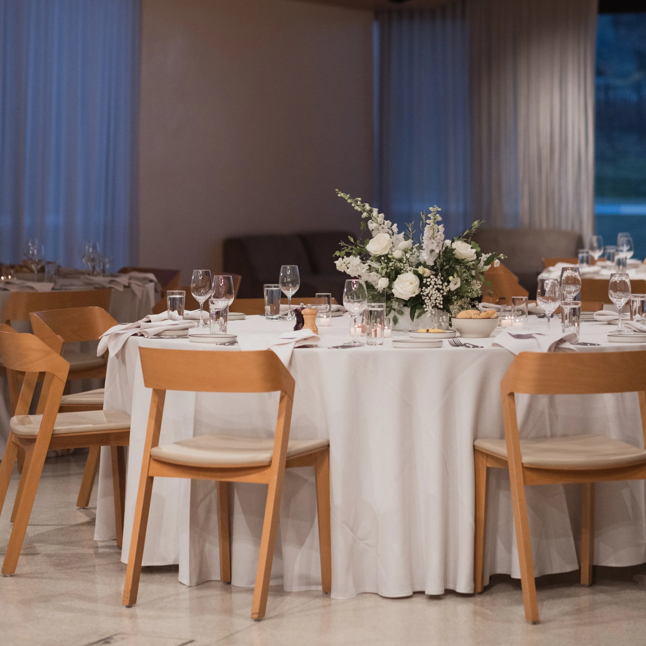 Elegant dining room with a round table set for a special event, featuring a white tablecloth, floral centerpiece with white roses, and neatly arranged glassware and napkins. Wooden chairs surround the table, and soft lighting creates a warm atmosphere.