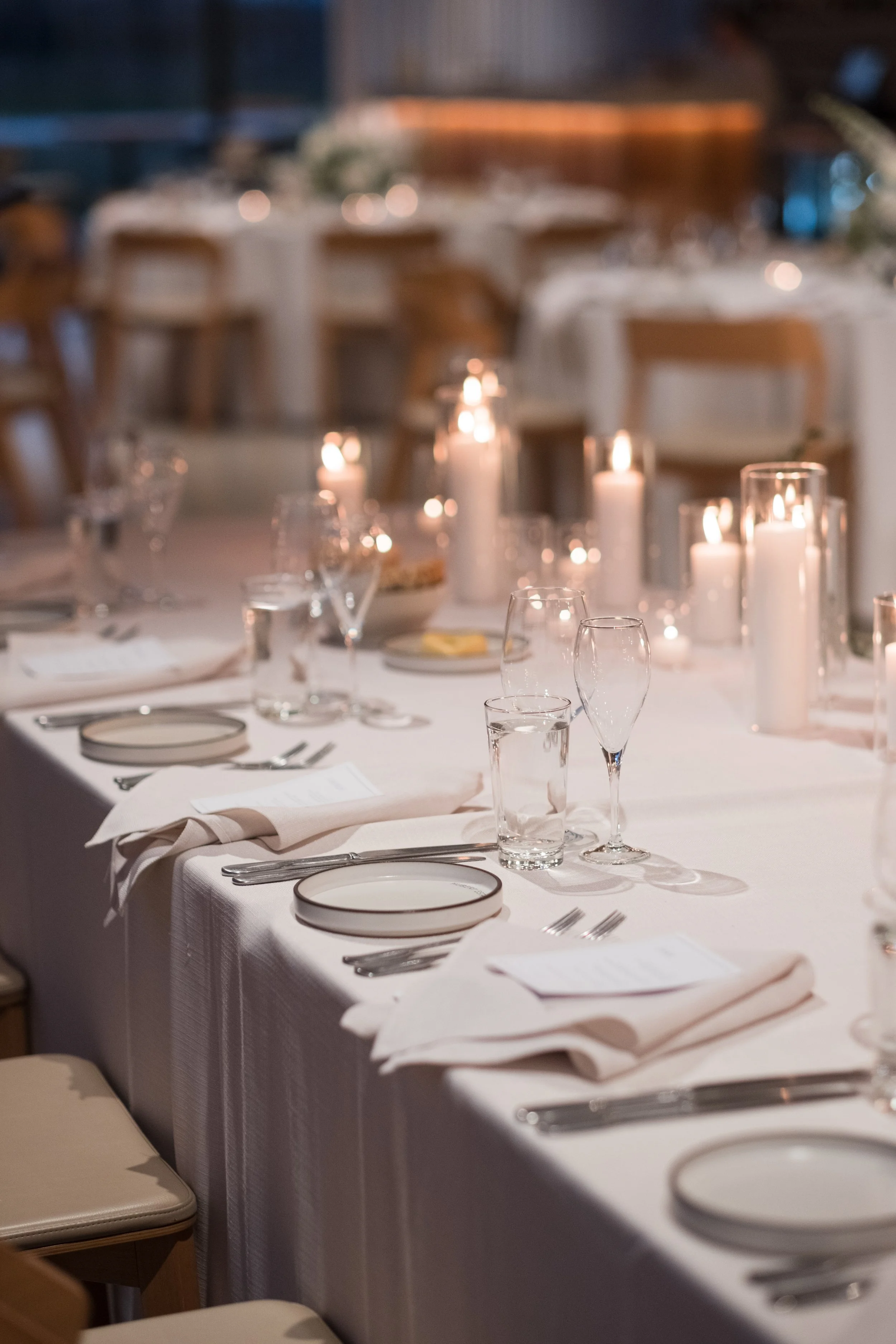 Elegant dinner table set with white tablecloth, plates, glasses, silverware, napkins, and lit candles in a dimly lit room, ready for a formal event.