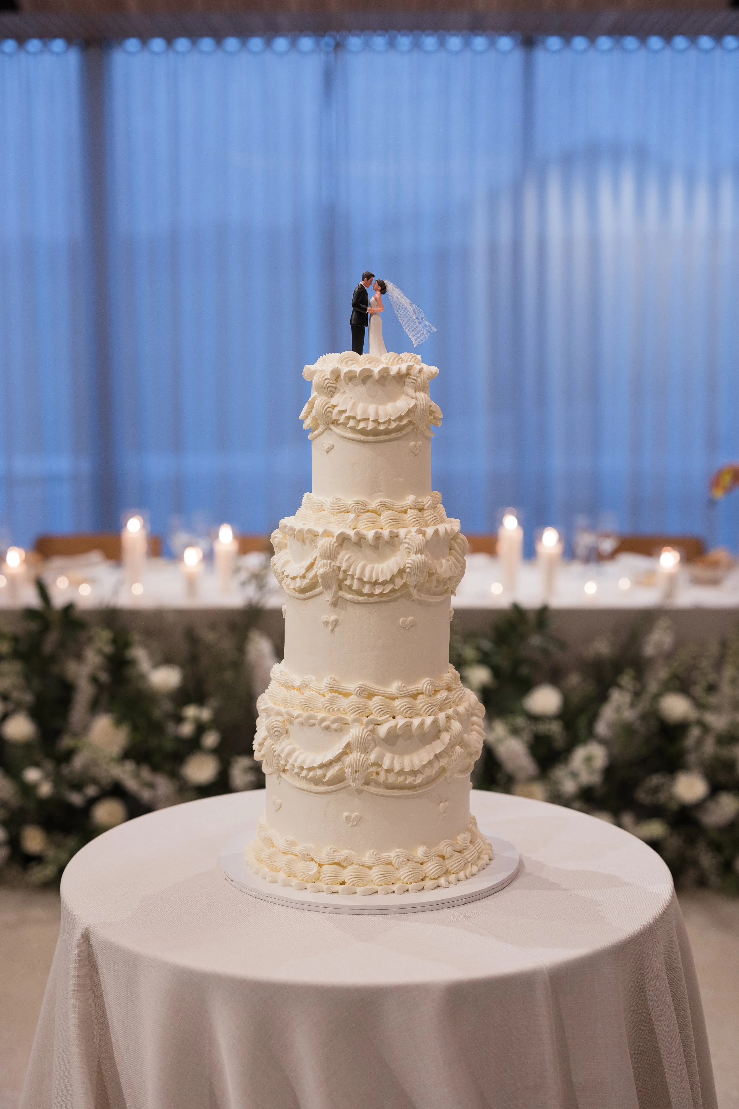 A tall, white wedding cake with intricate piping decoration, topped with a miniature bride and groom figurine, placed on a round table with a white tablecloth in a decorated reception hall.