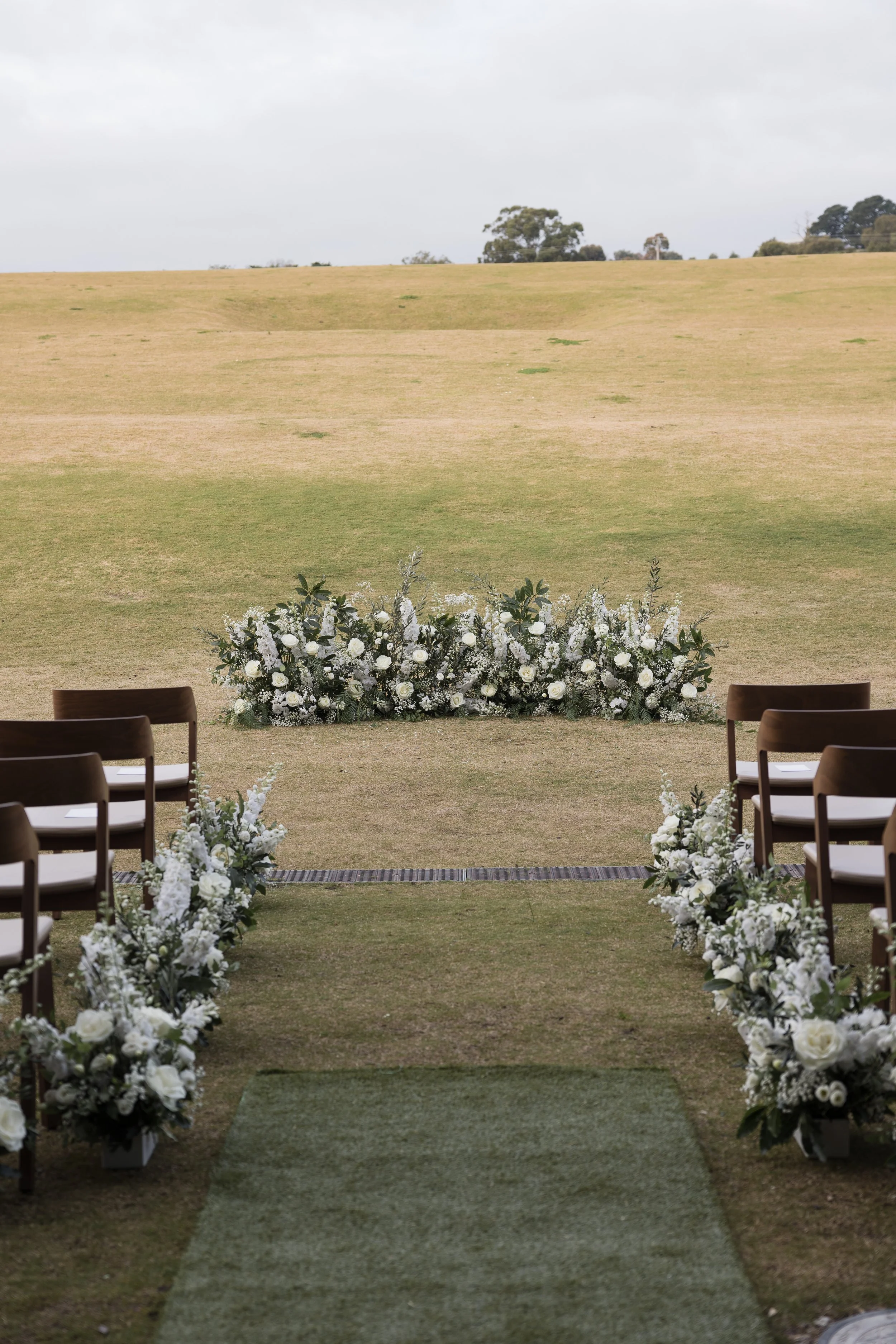 Outdoor wedding ceremony setup on grass with white floral arrangements along the aisle and at the altar, overlooking a field and trees.