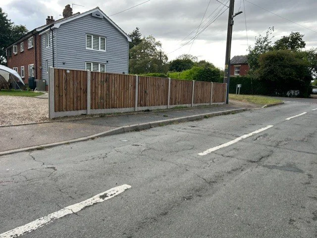A wooden fence along the sidewalk next to a street, with a two-story house and a tent visible behind the fence under a cloudy sky.