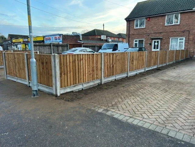 A new wooden fence installed along a sidewalk, with a brick house and parked cars in the background.
