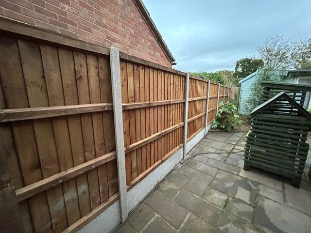 Newly installed wooden fence along a brick house with a paved backyard, shed, and some plants.