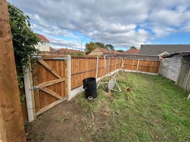 View of a backyard with a newly installed wooden fence, a garden gate, a wheelbarrow, a black trash bin, and a cloudy sky.